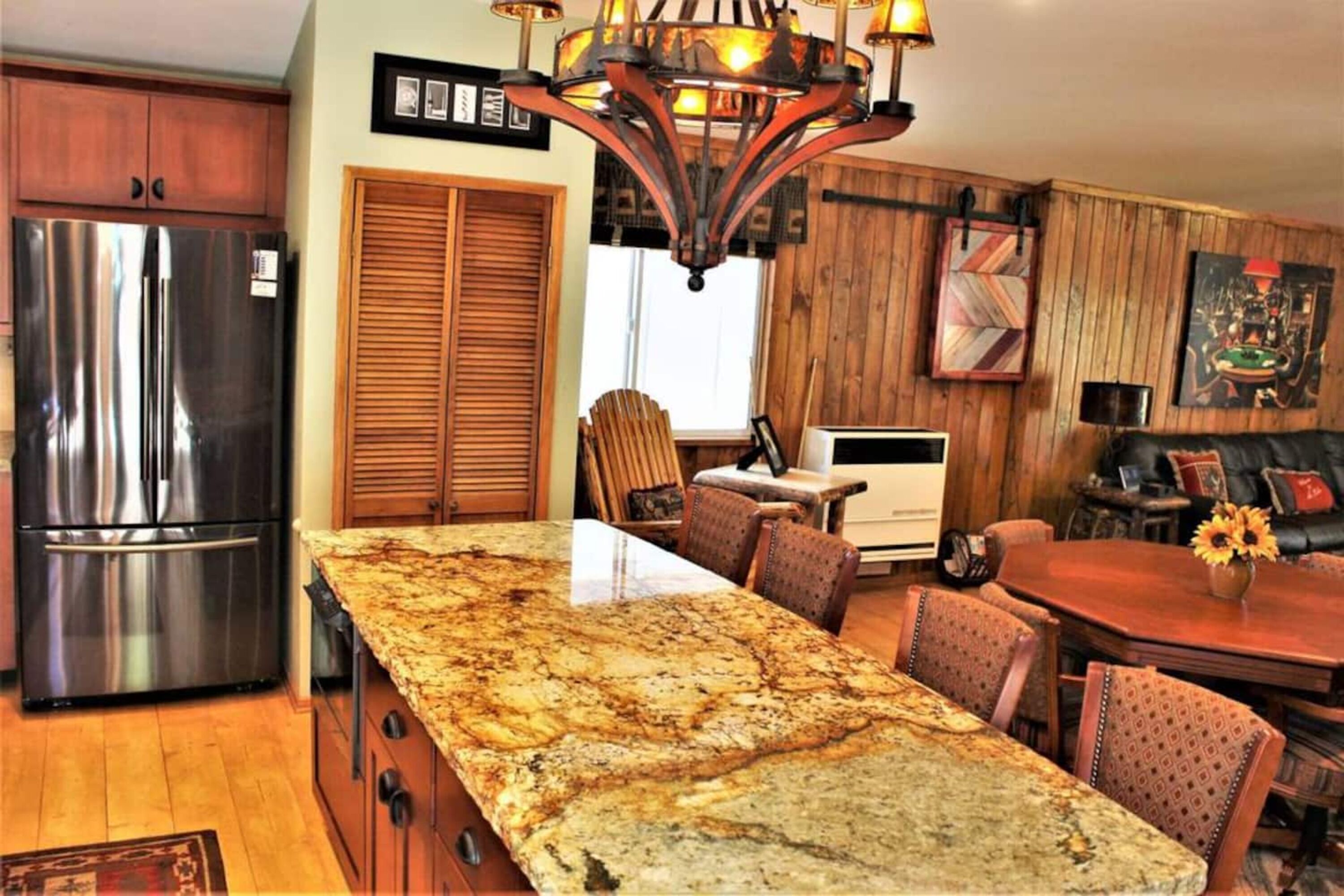 Una cocina con una isla de granito, nevera de acero inoxidable, armarios de madera y una araña.