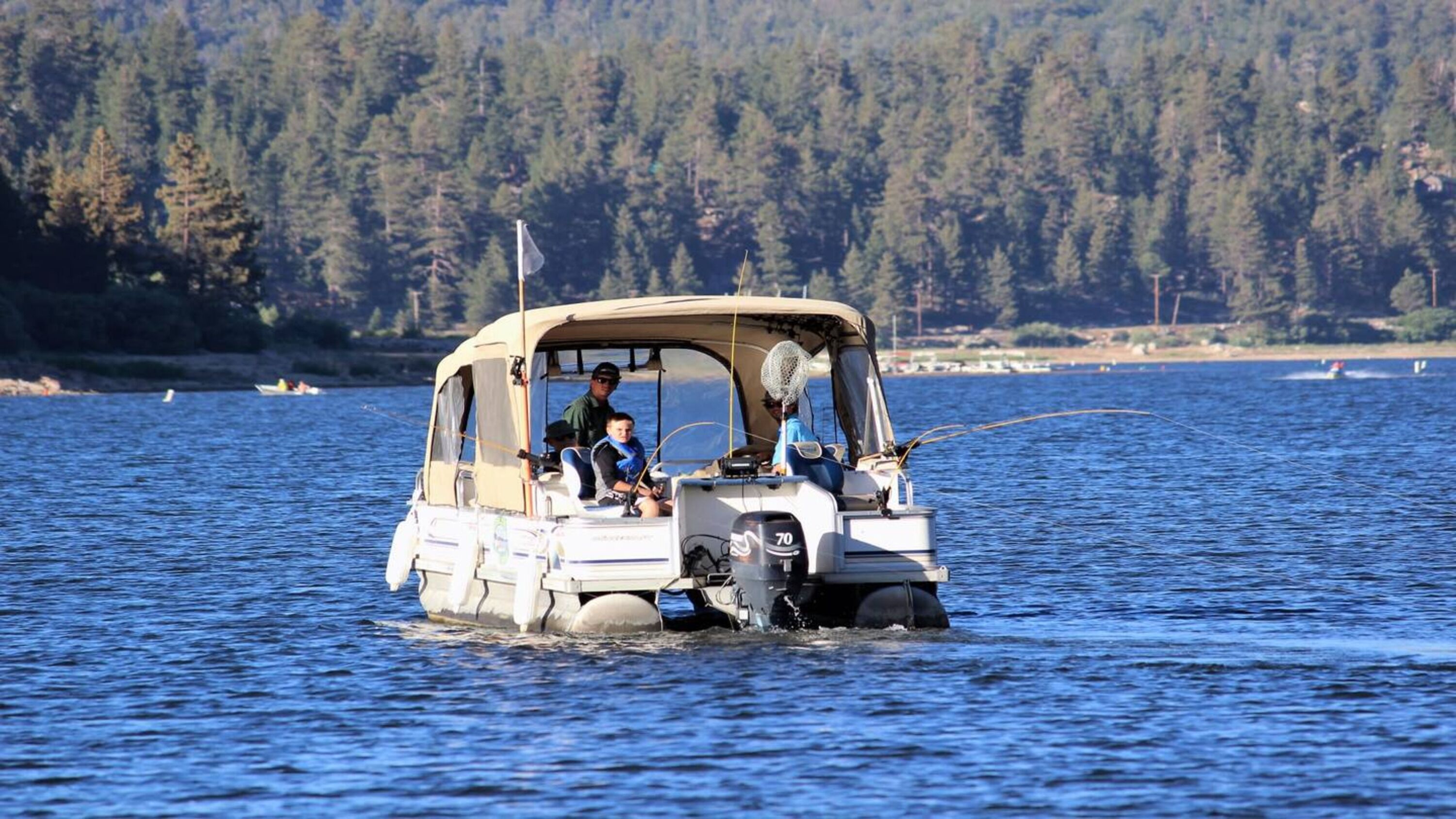 A small boat with two people on board, moving through a body of water with a forested shoreline in the background.