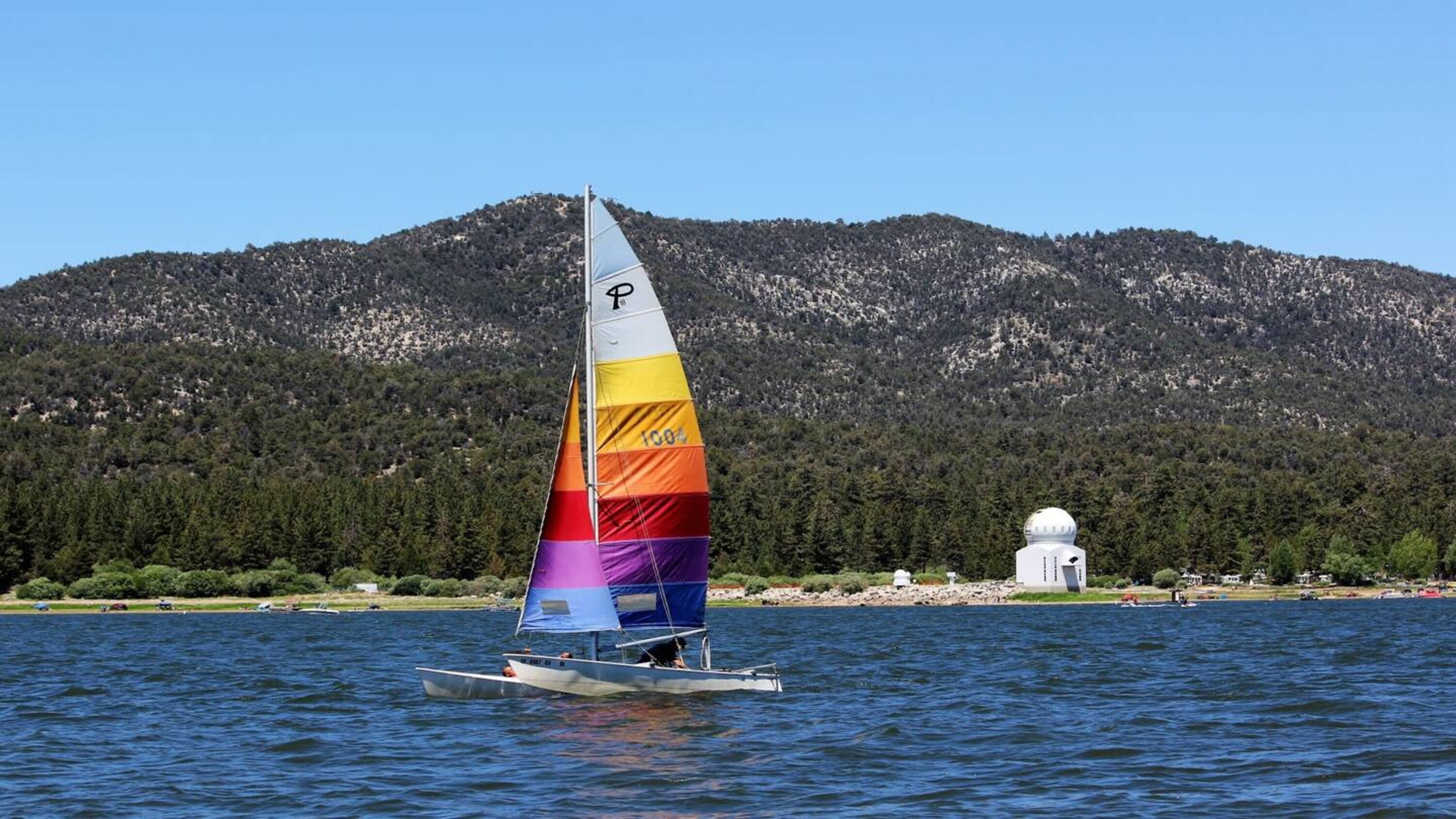 Un velero con una vela colorida en un lago, con una montaña boscosa al fondo.