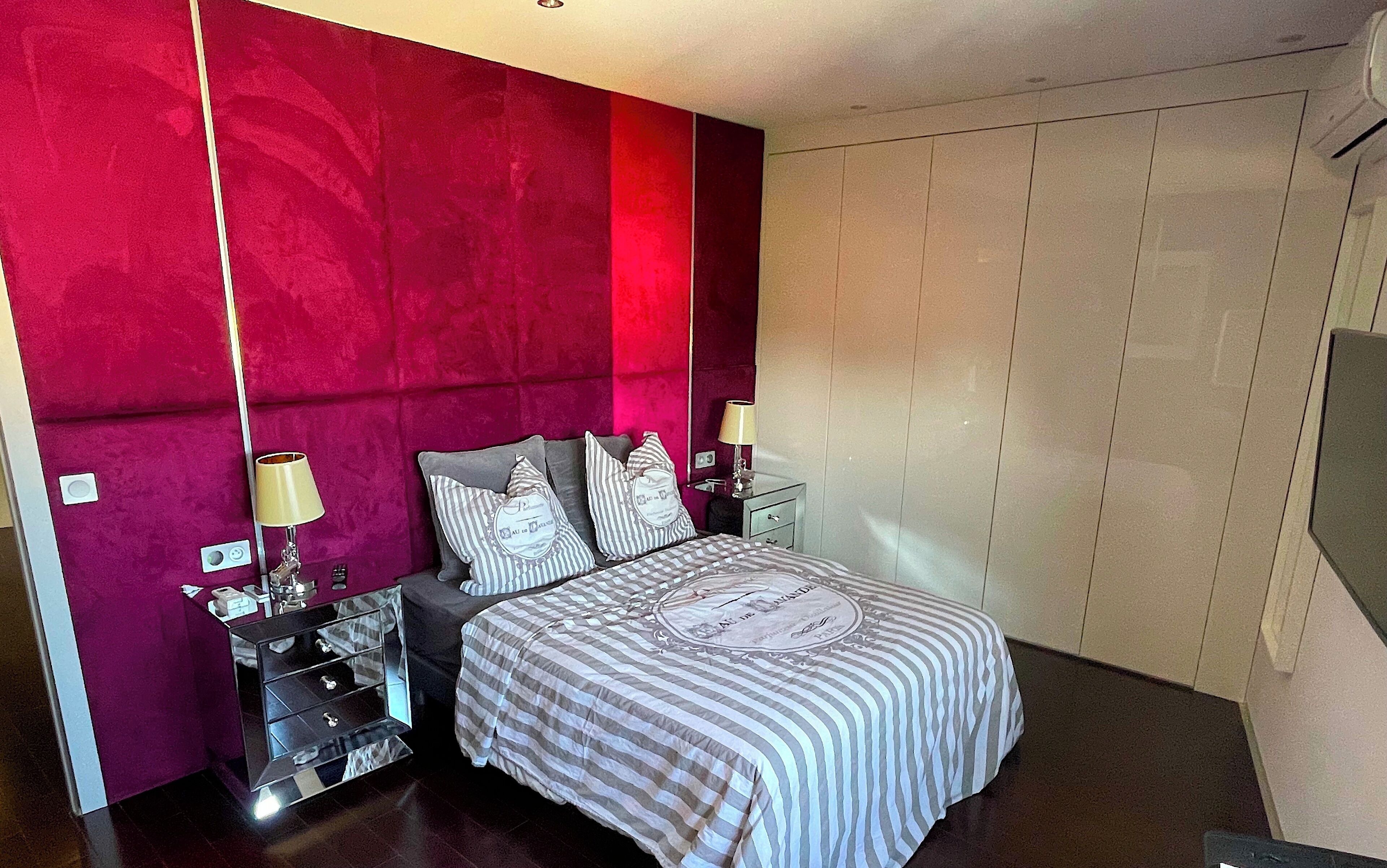 A bedroom with a red accent wall, a bed with striped bedding, two bedside tables with lamps, and a wardrobe.