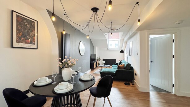 A modern dining area with a round table, black chairs, and a vase of flowers. There is a sofa and a television in the background. - 2 Bedroom Queen Street (Newcastle-upon-Tyne)