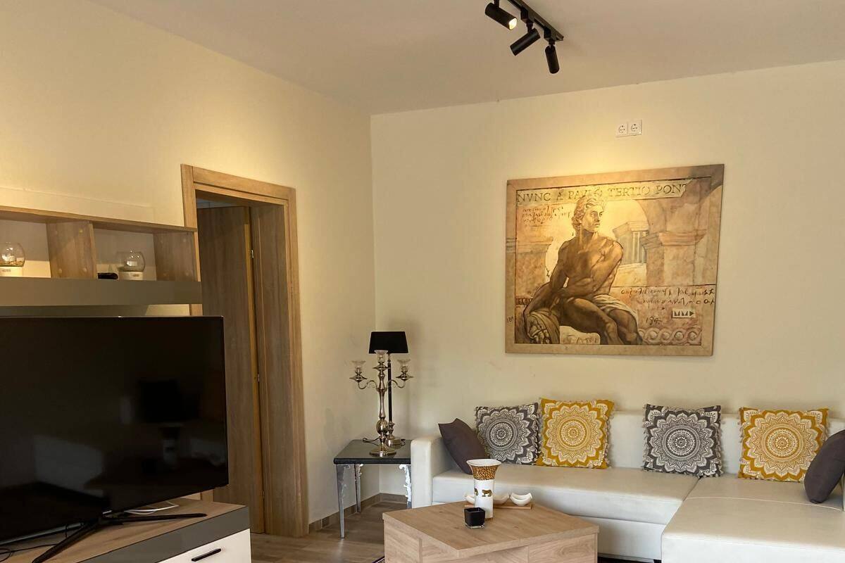 A modern living room with a flat-screen TV, a white sofa, patterned cushions, and a large wall painting.