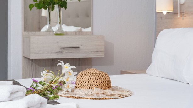 A neatly arranged hotel room with a bed, a bedside table with a woven basket, a vase with flowers, and a mirror above a wooden dresser. - Alba Marea (Dio-Olympos)