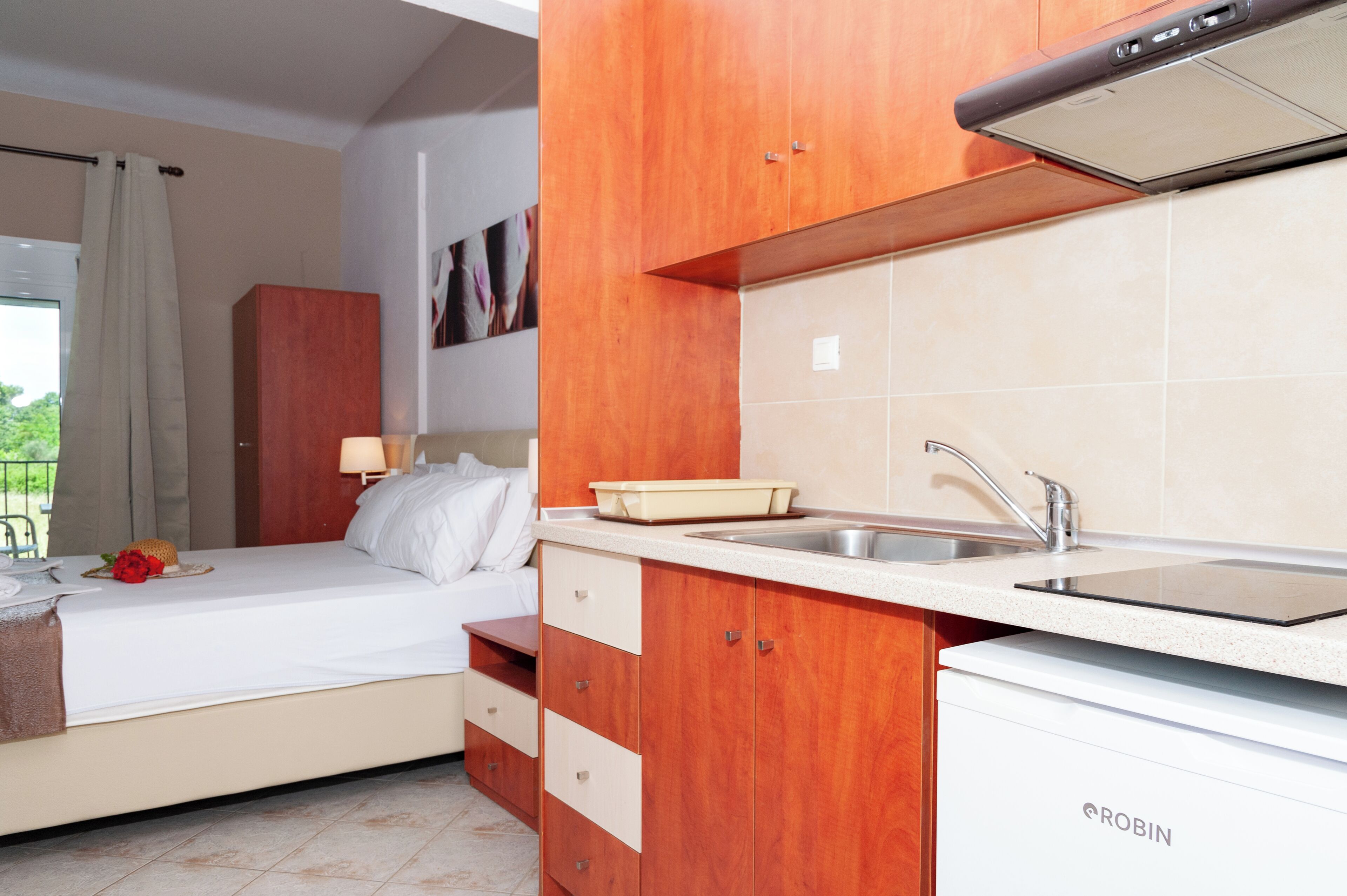 A compact hotel room with a kitchenette, a bed with white linens, and a view of greenery outside.
