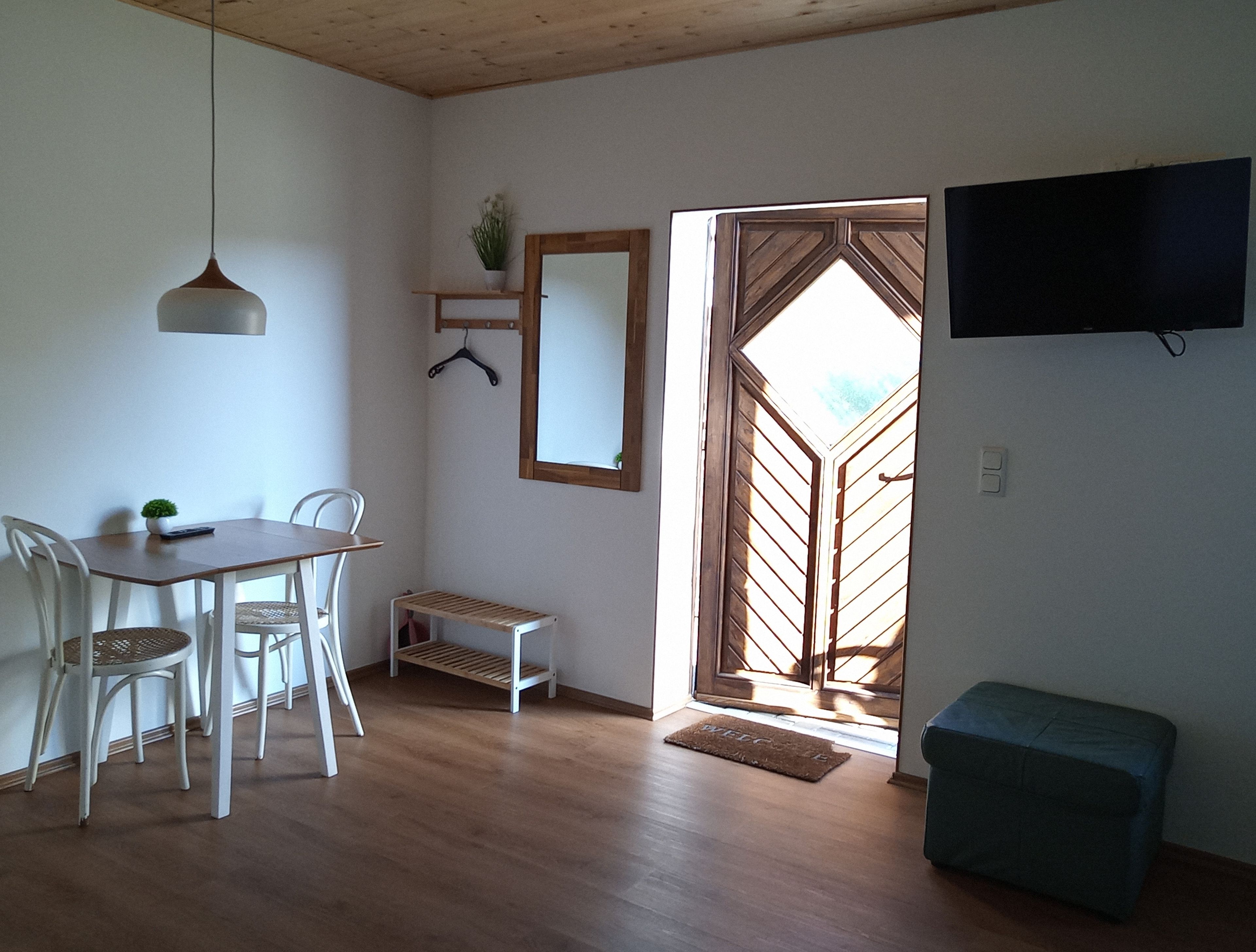 Una habitación con una mesa de comedor de madera, sillas blancas, una lámpara colgante, una planta pequeña, un espejo y un televisor en la pared.