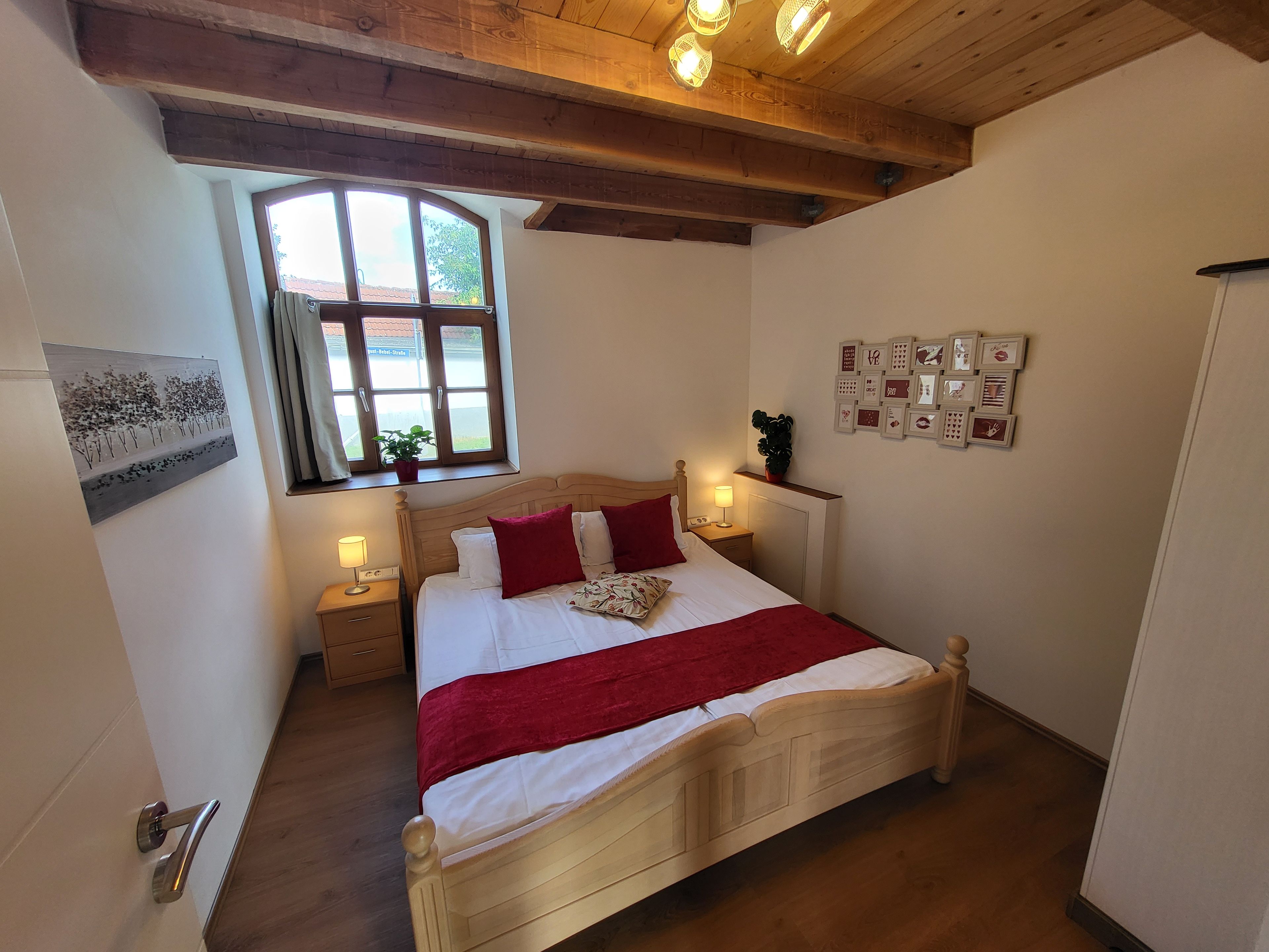 A bedroom with a wooden bed, a window with curtains, bedside lamps, and a wall with framed pictures.