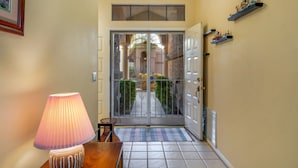 A hallway with a tiled floor, a wooden table with a lamp, a door with a metal gate, and a wall-mounted shelf with decorative items.