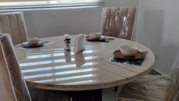 Ett runt matbord med fyra stolar, var och en täckt med skyddande plast. En flaska handsprit och en hållare för pappersnäsdukar står på bordet.