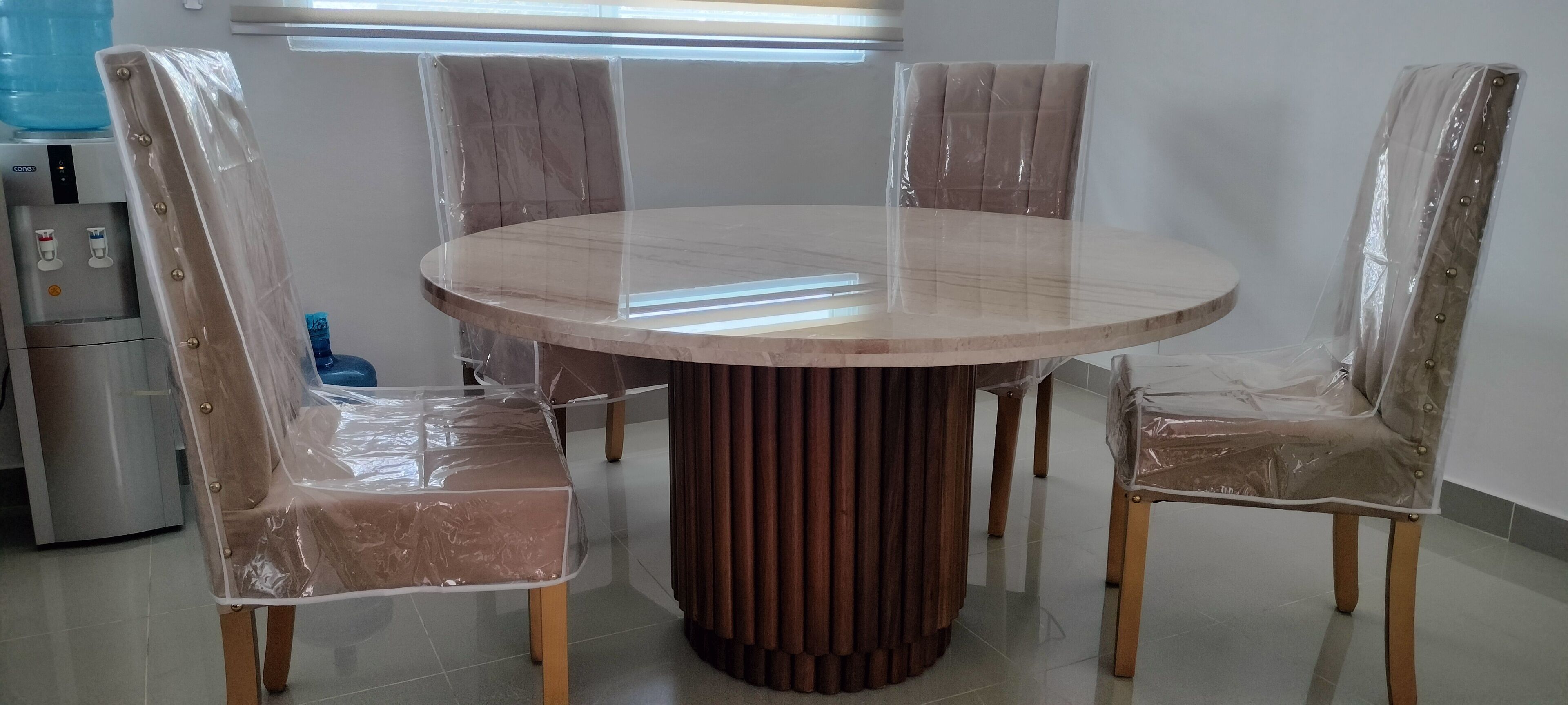 Ein runder Esstisch mit vier Stühlen, jeweils mit Plastikfolie abgedeckt. Im Hintergrund sind ein Wasserspender und ein Kühlschrank zu sehen.