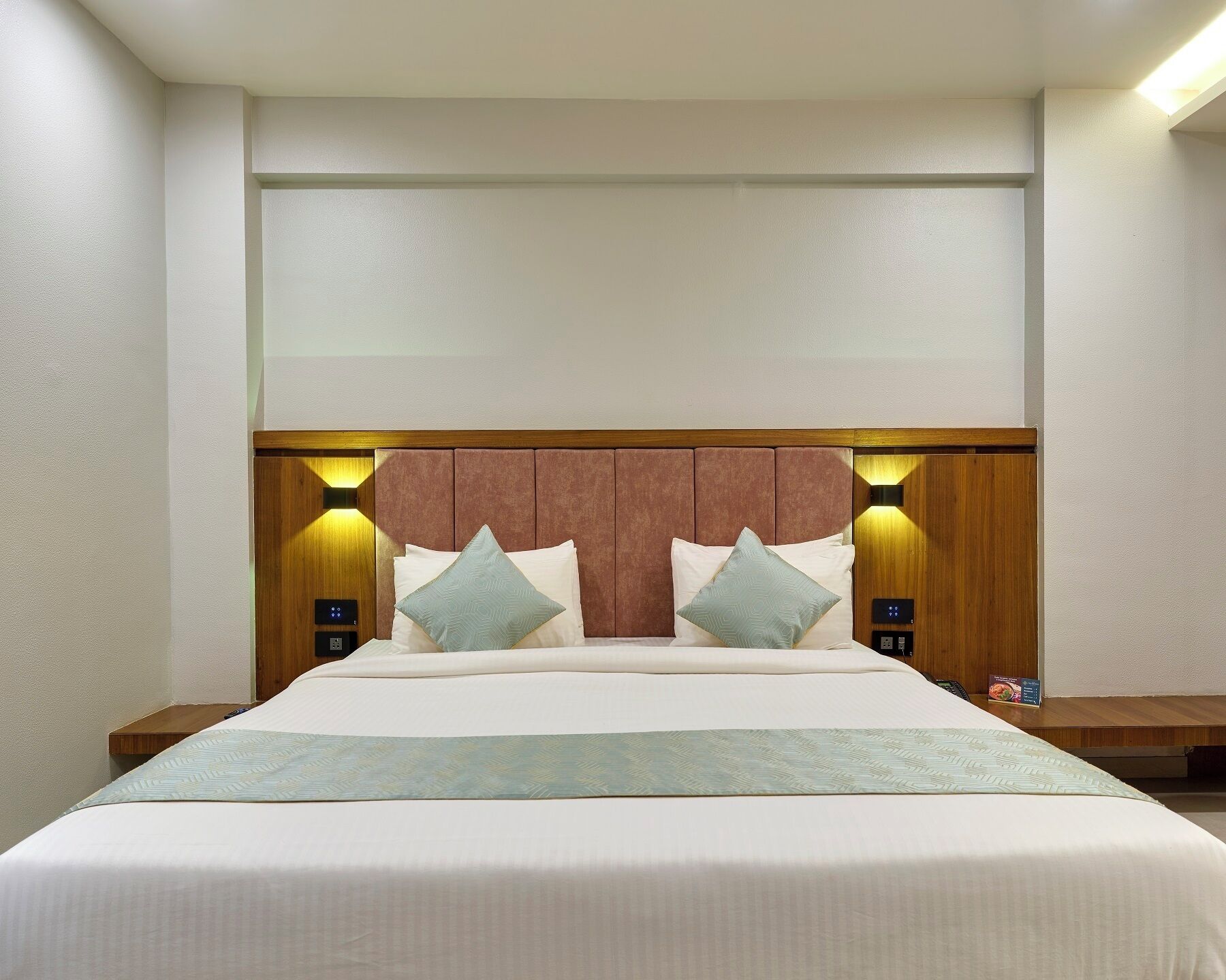 Habitación de hotel con una cama grande, cabecero de madera, dos mesitas de noche e iluminación empotrada.