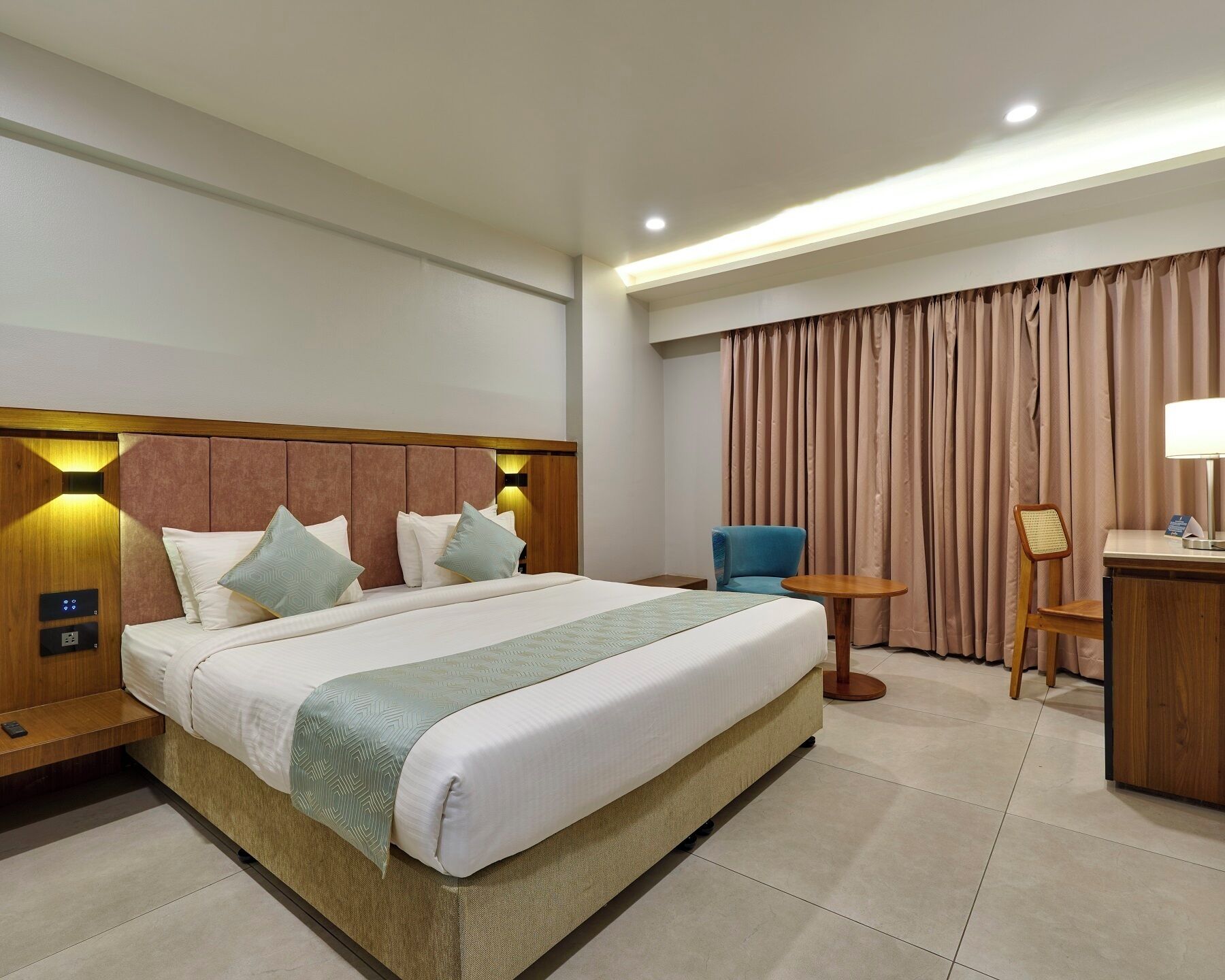 A hotel room with a large bed, bedside tables, a desk, a chair, and a curtain.