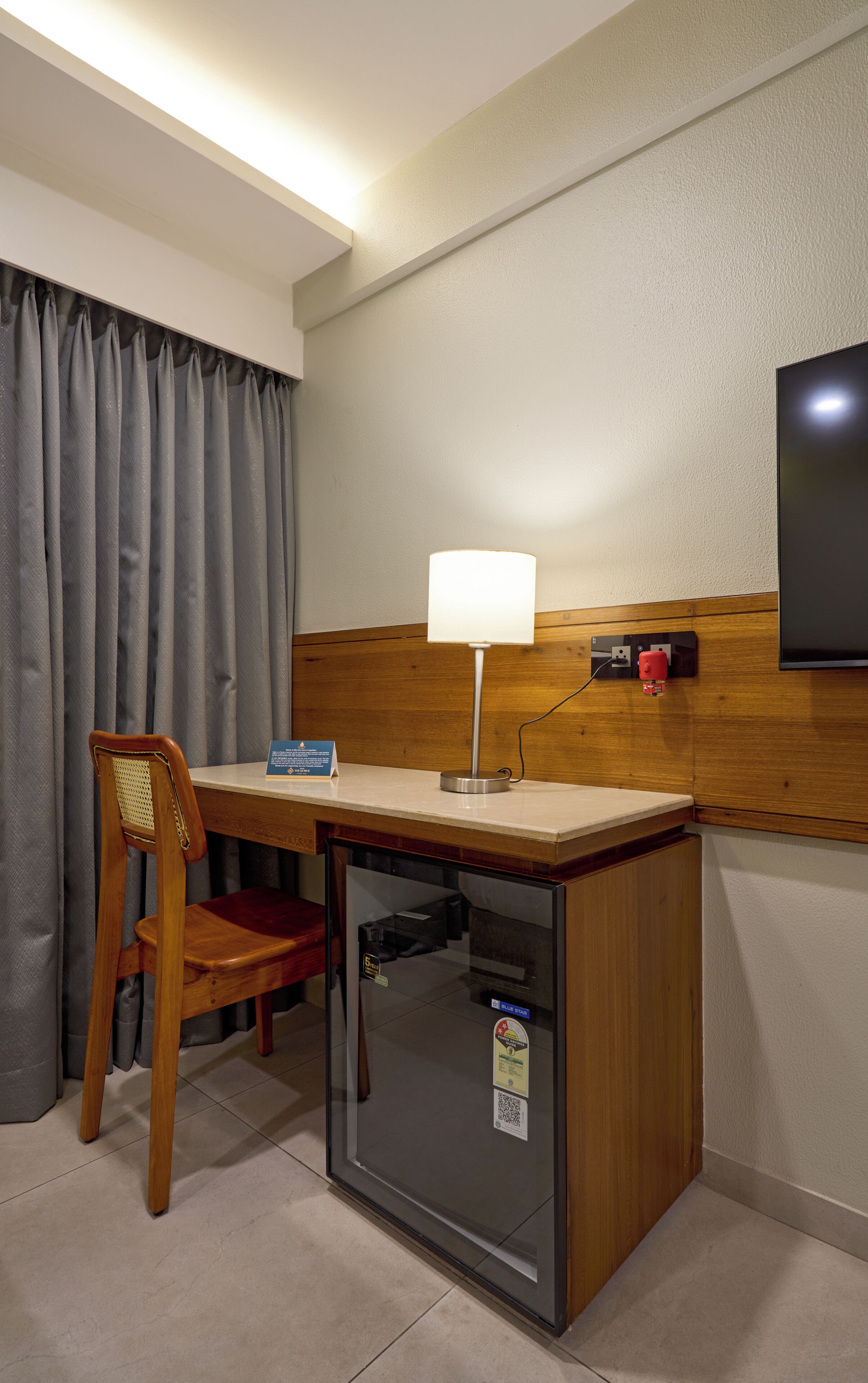 A compact hotel room with a wooden desk, a chair, a small refrigerator, a flat-screen TV mounted on the wall, and a window with curtains.