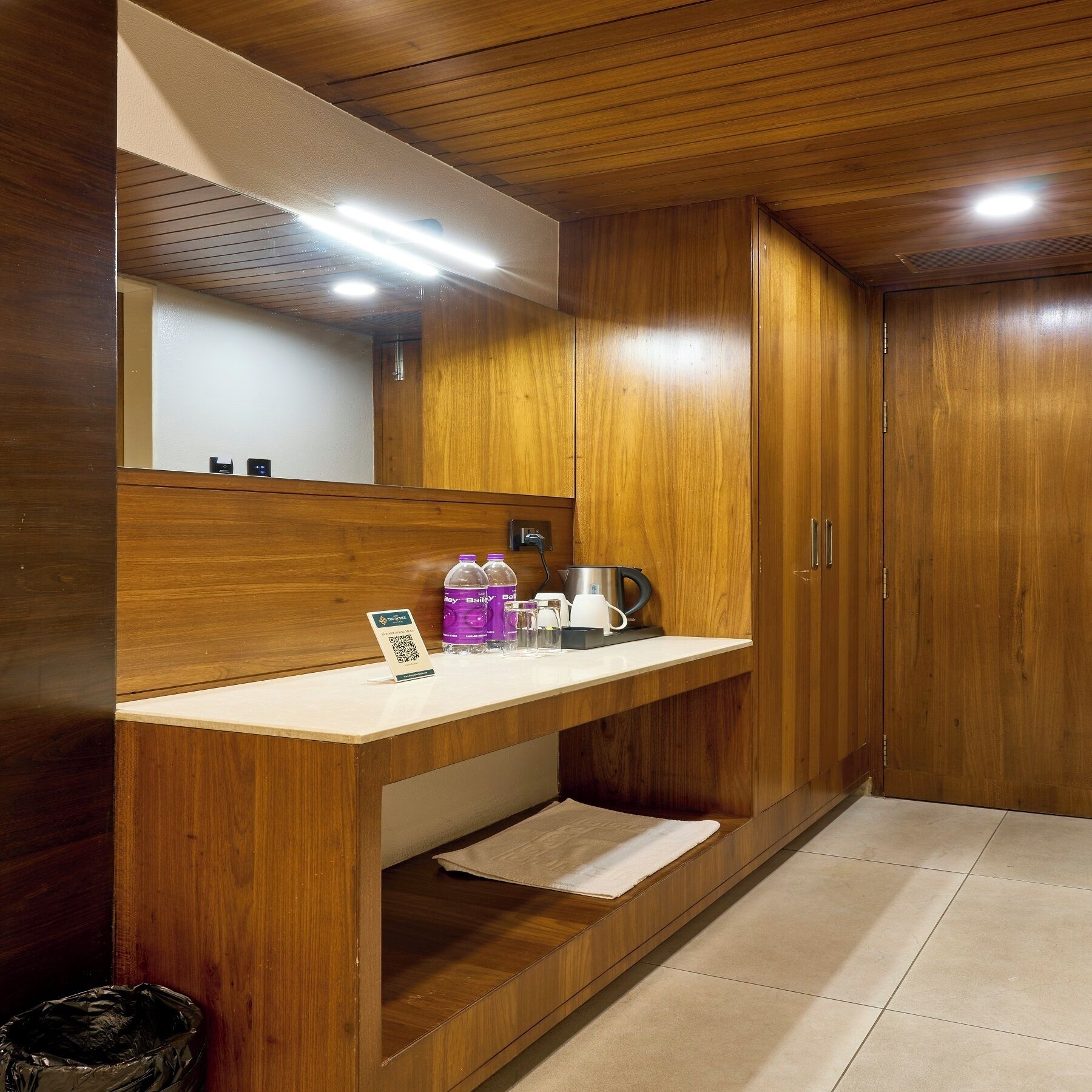 Une chambre d’hôtel équipée d’une coiffeuse en bois, d’un miroir, d’un lavabo et d’un petit plan de travail sur lequel se trouvent une bouilloire, des tasses et un panneau avec un code QR.