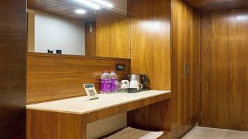Une chambre d’hôtel équipée d’une coiffeuse en bois, d’un miroir, d’un lavabo et d’un petit plan de travail sur lequel se trouvent une bouilloire, des tasses et un panneau avec un code QR.