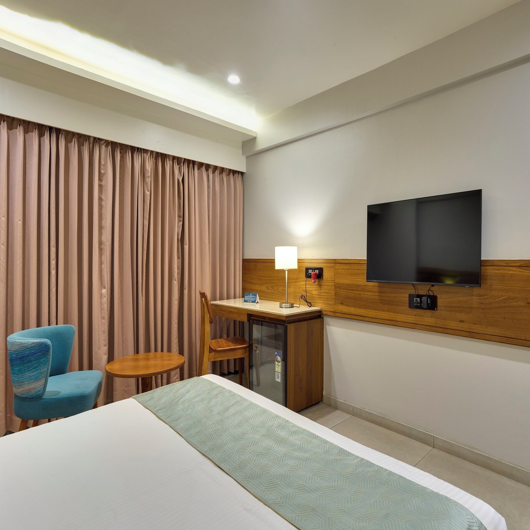 A hotel room with a bed, a desk with a lamp, a television, a chair, and a small table.