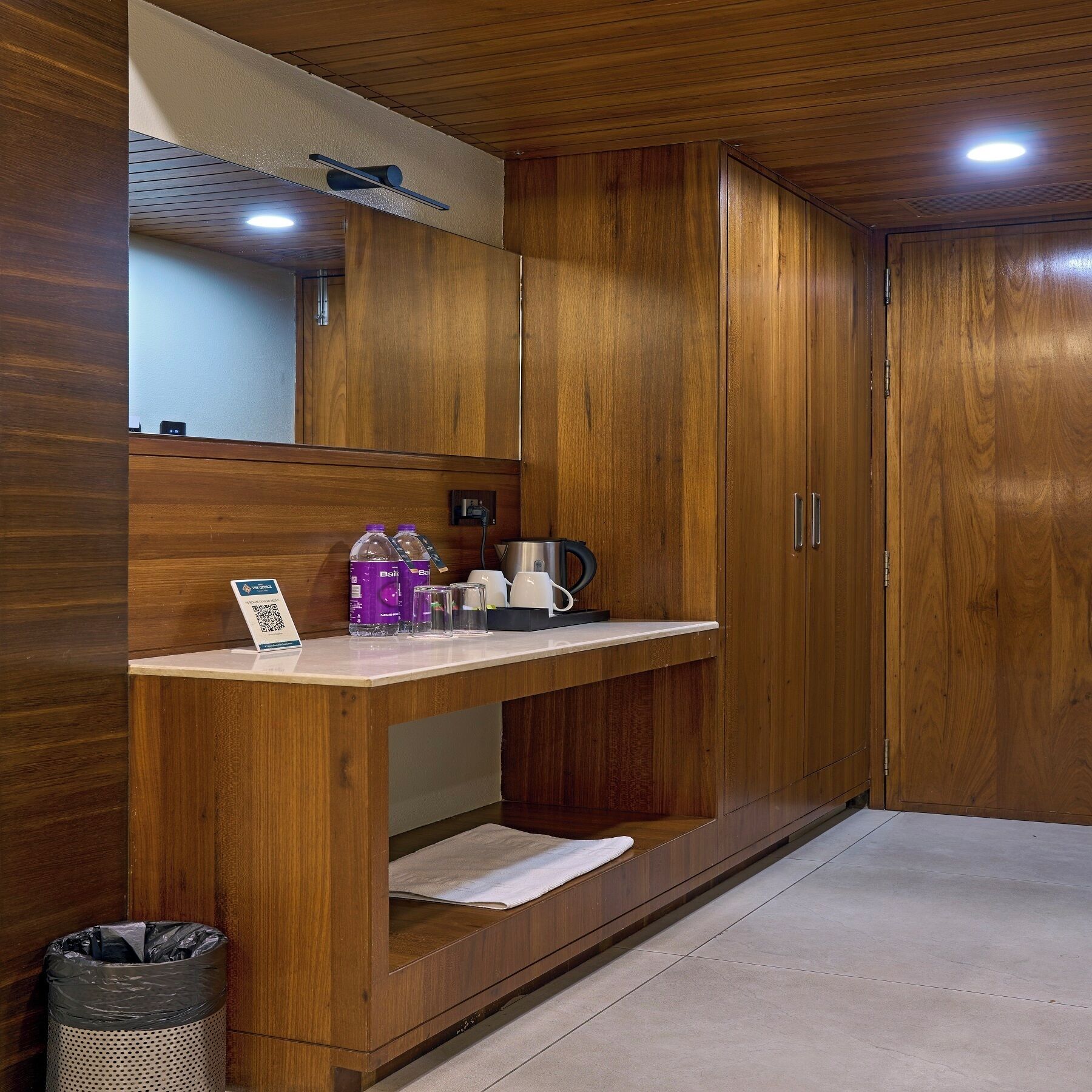 A wooden cabinet with a mirror, a small countertop with a coffee maker, and a trash can.