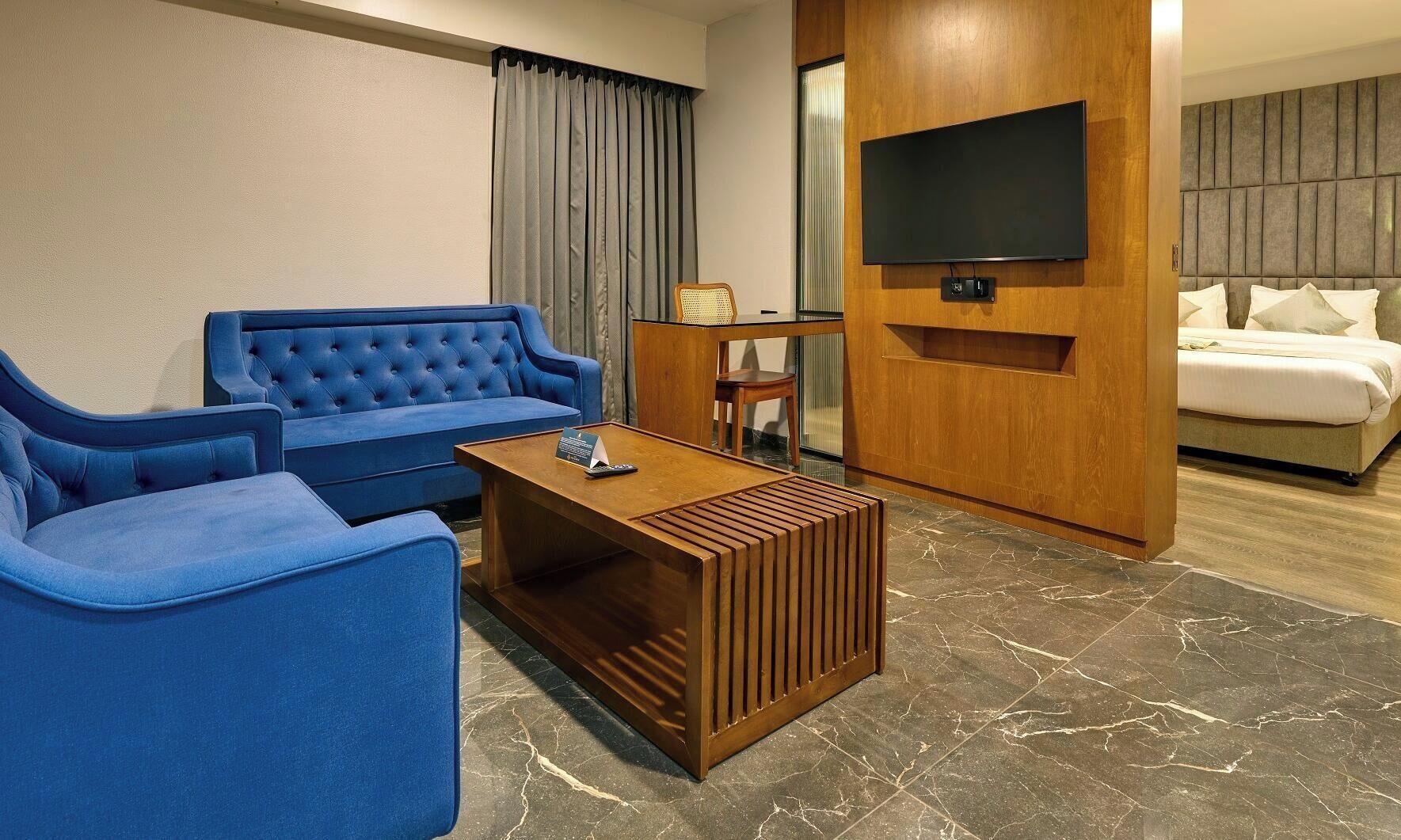 A hotel room with a blue sofa set, a wooden coffee table, a TV mounted on a wooden panel, and a bed with white bedding.