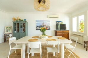 Une salle à manger avec une table blanche, des chaises, un vase de fleurs, un buffet vert et un tableau représentant un paysage urbain.