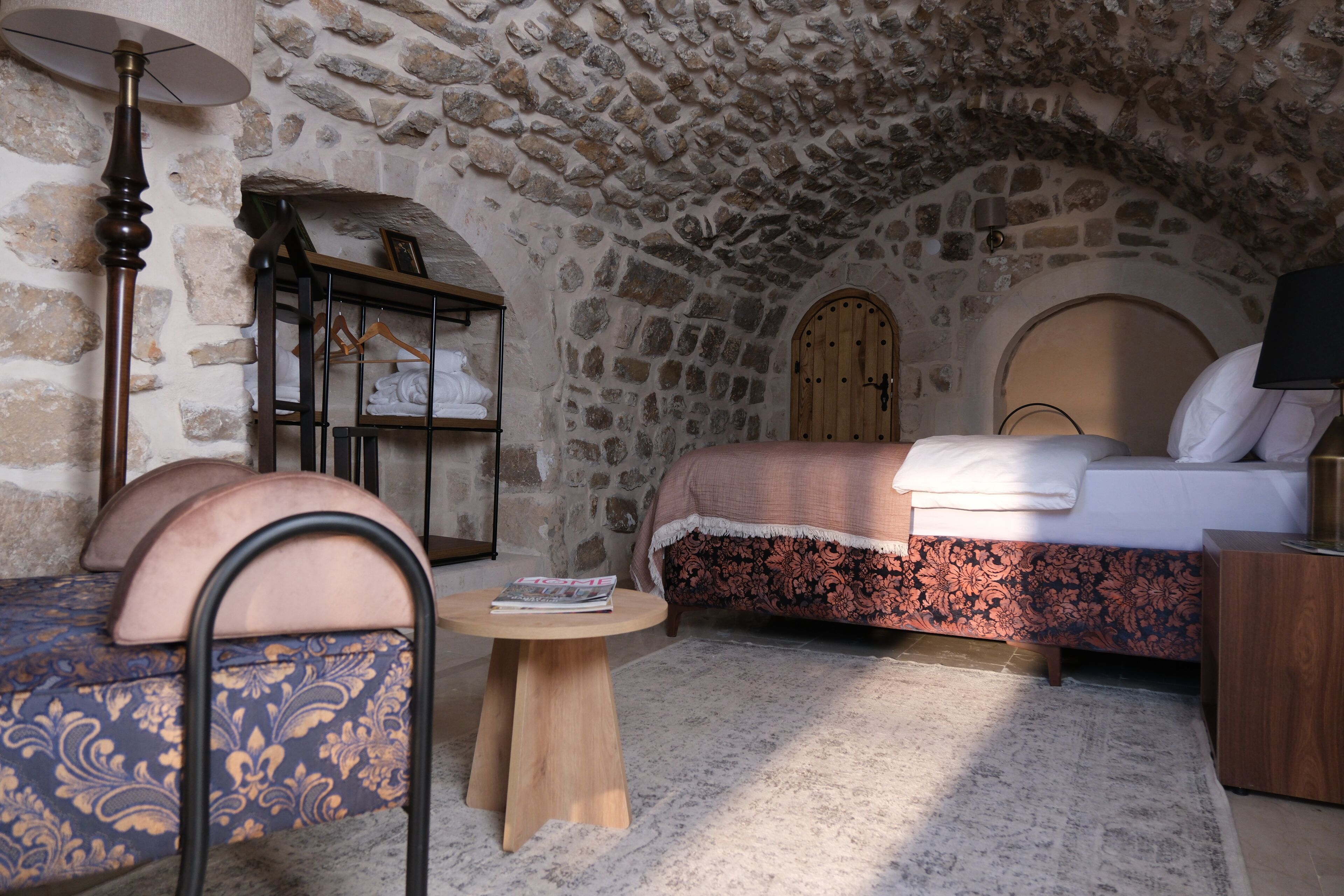 A cozy room with a stone ceiling, a bed with a floral bedspread, a round wooden table, and a chair with a patterned cushion.
