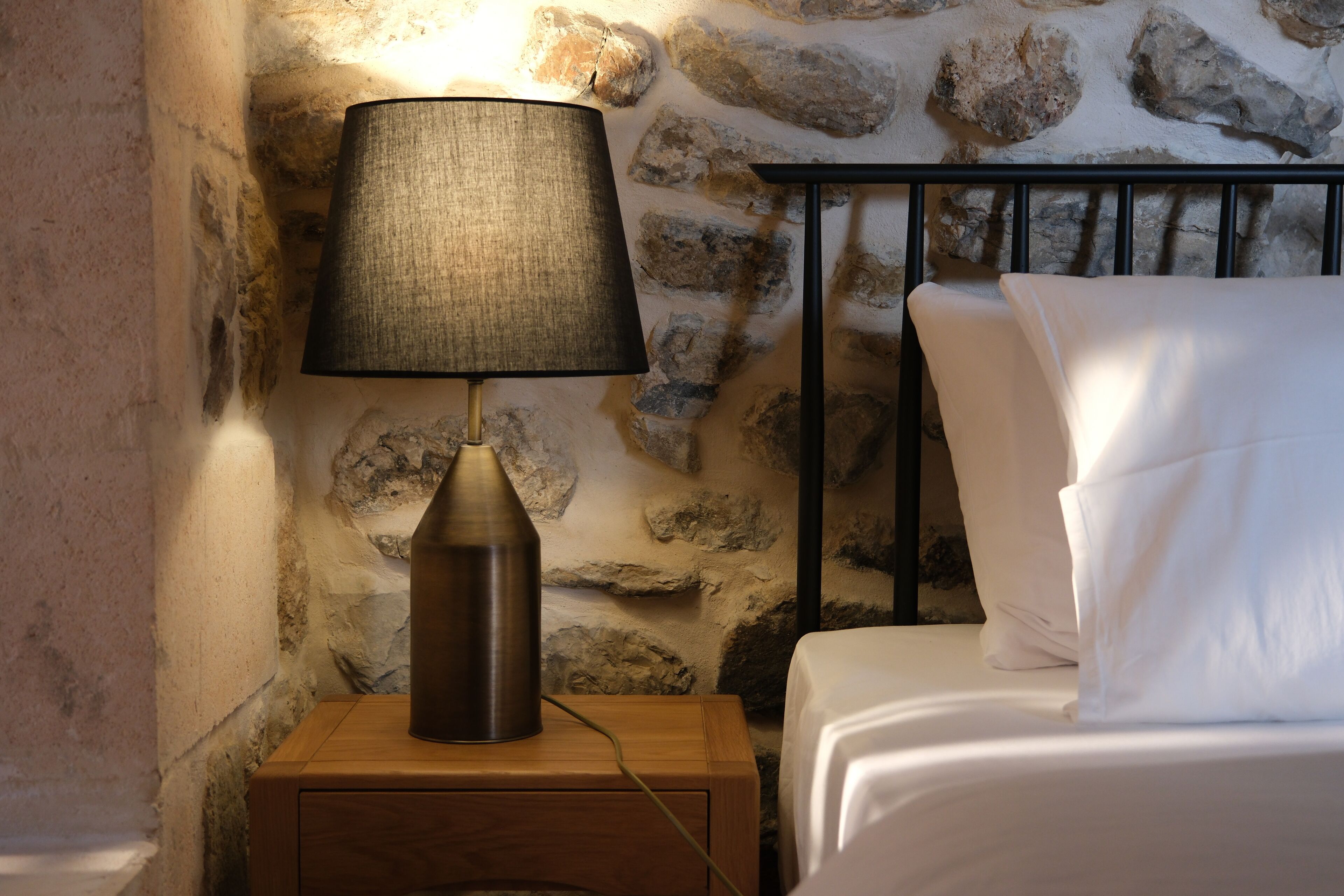 A bed with white linens, a bedside lamp with a dark shade, and a stone wall backdrop.
