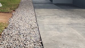 Ein Betonweg, der von einem Kiesbett gesäumt wird, führt zu einem Gebäude mit einer Backsteinmauer. Eine Leiter lehnt an einem Baum.