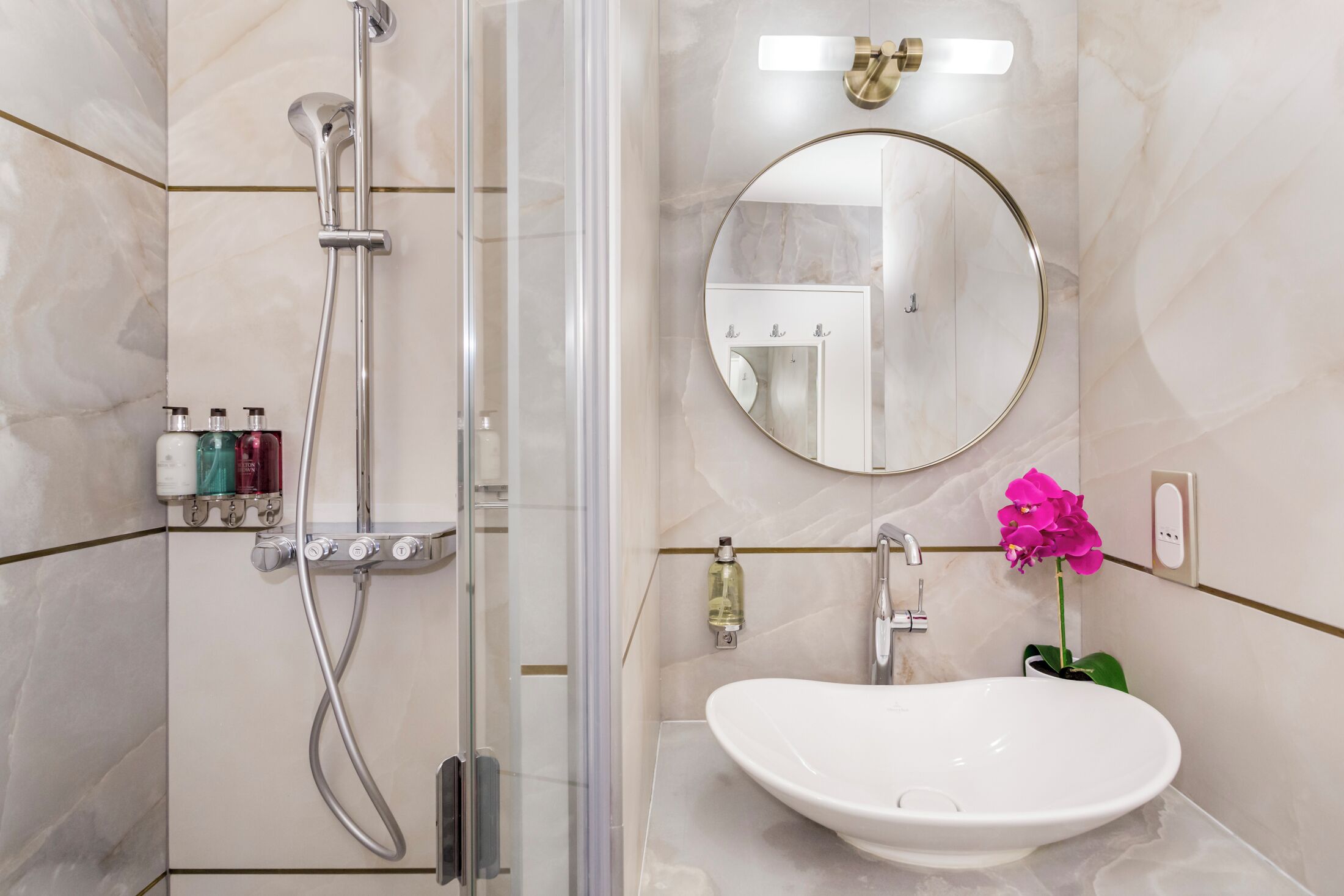 Un baño moderno con ducha envuelta en vidrio, un espejo redondo, un lavabo y artículos de aseo en la pared.