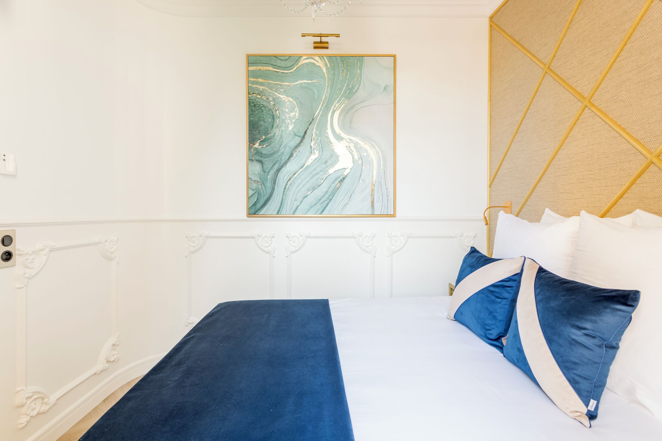 A neatly made bed with a blue bedspread and matching pillows, a marble artwork above it, and a decorative headboard.