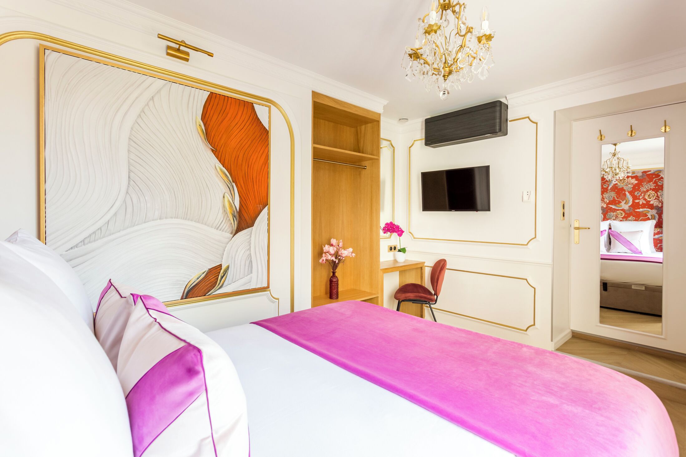 A modern hotel room with a large bed, a TV mounted on the wall, a wooden wardrobe, and a small desk with a chair.