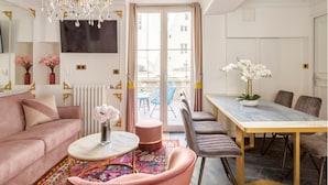A modern living room with a pink sofa, a round coffee table, a chandelier, and a dining area with a table and chairs. - Merveil Luxury Suites - Bac I-II (Paris)