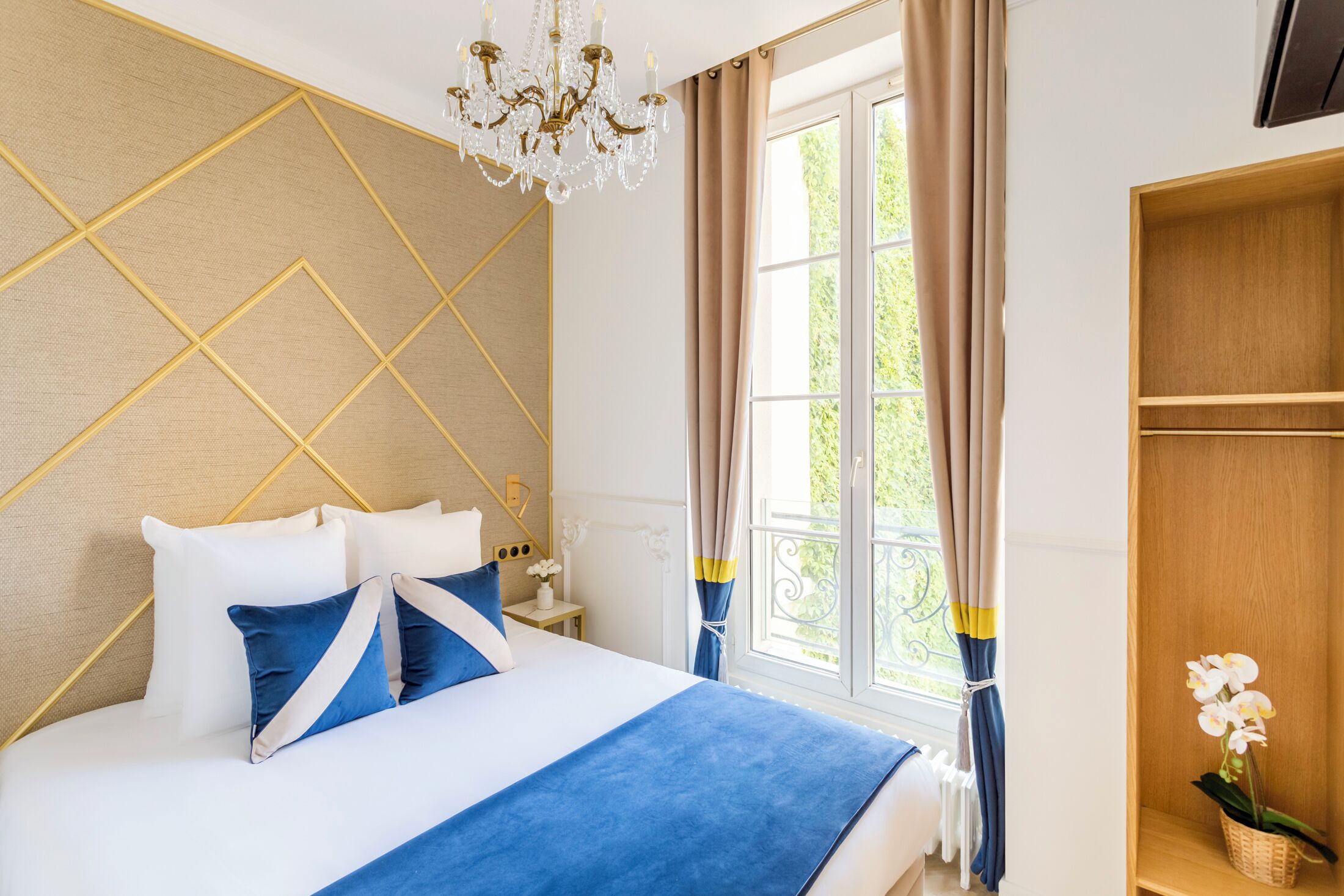 Une chambre à coucher avec un lit, un lustre, une fenêtre avec des rideaux et une étagère en bois ornée d’une orchidée.
