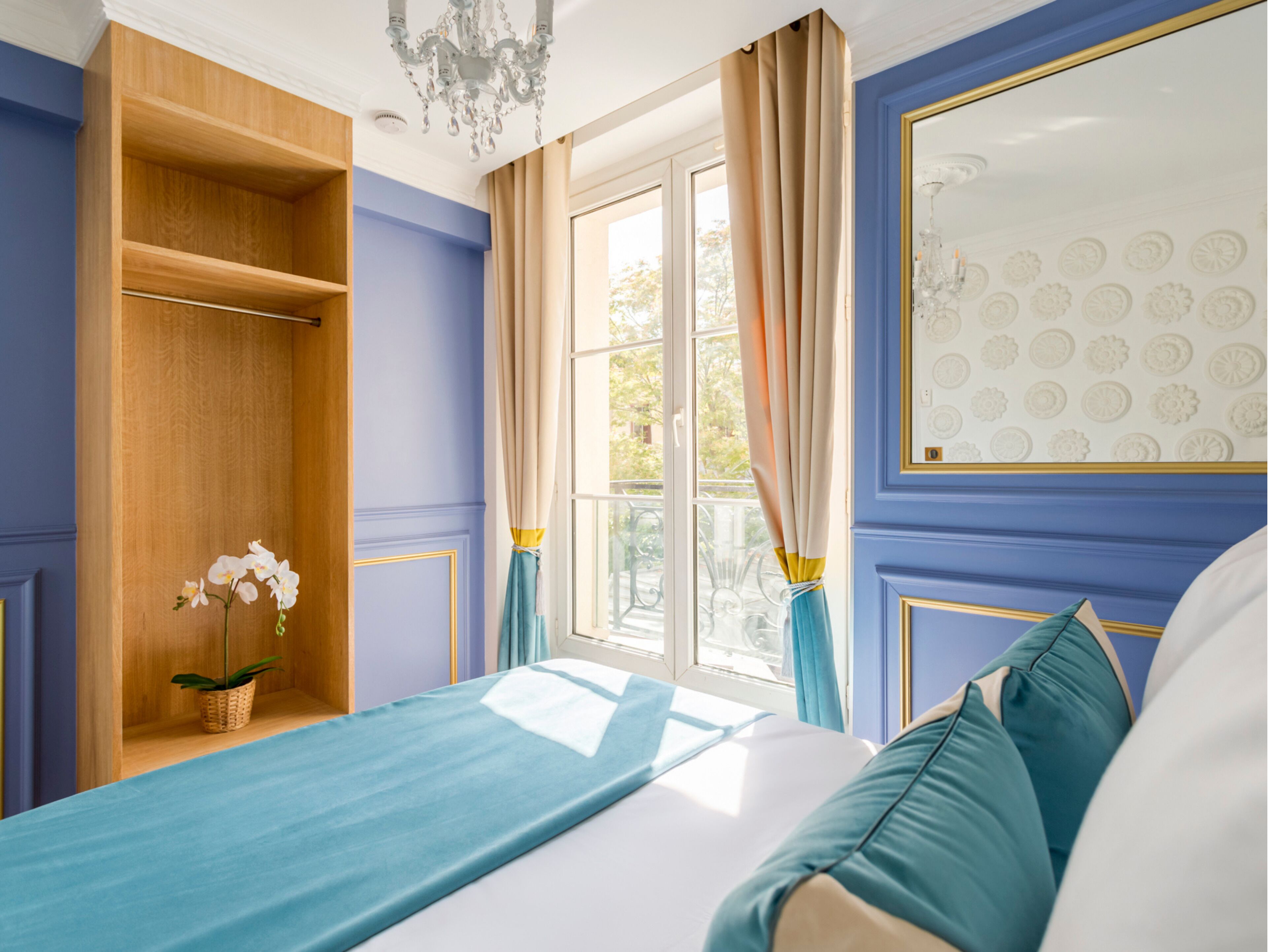 A bedroom with a large window, a wooden wardrobe, a chandelier, and a bed with blue bedding.