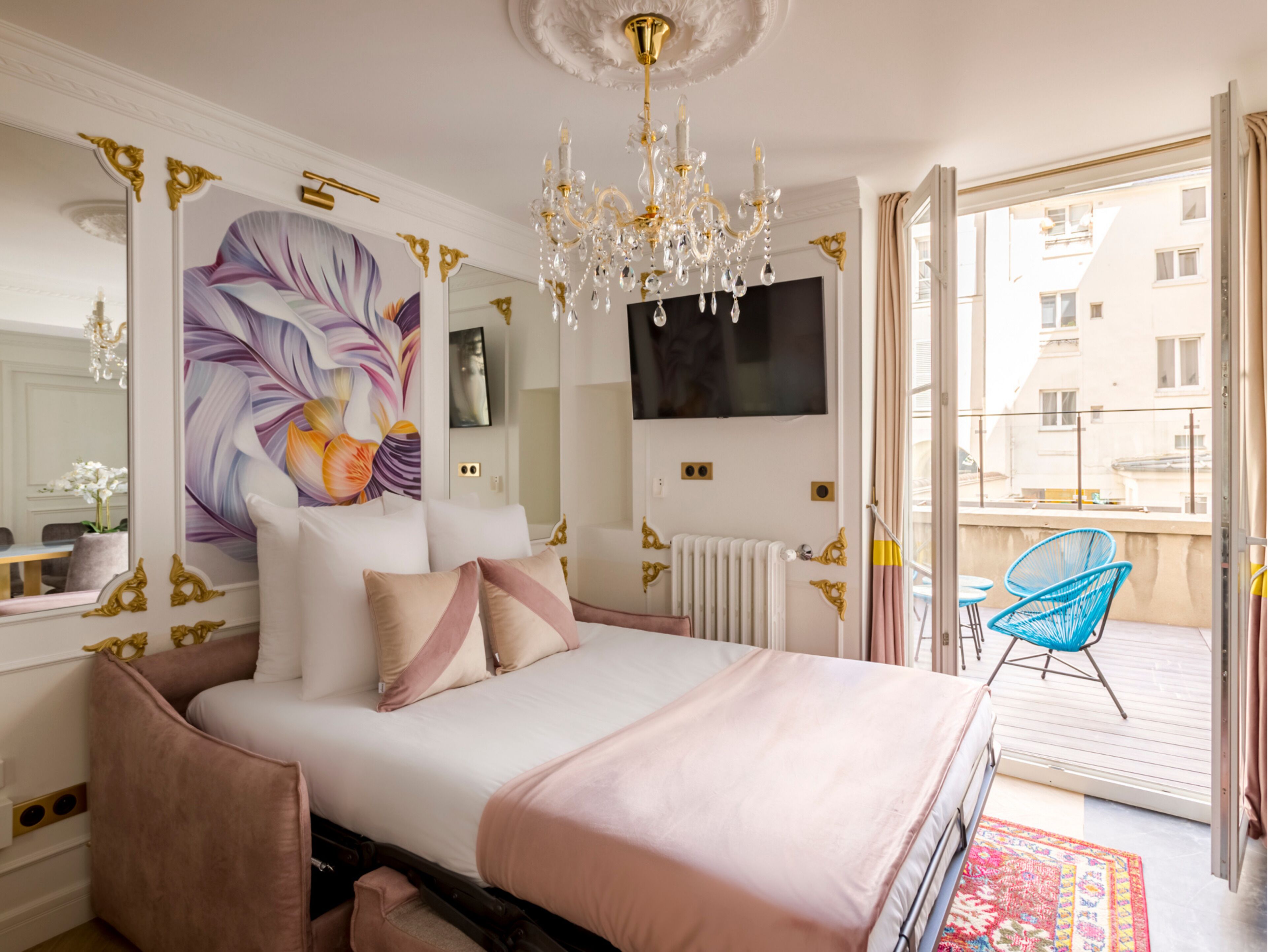 A bedroom with a large bed, a chandelier, a TV, and a balcony with chairs.