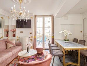A modern living room with a pink sofa, a round coffee table, a chandelier, and a dining area with a table and chairs. - Merveil Luxury Suites - Bac I-II (Paris)