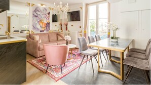 A modern kitchen with a dining area, featuring a pink sofa, a chandelier, and a large window with curtains. - Merveil Luxury Suites - Bac I-II (Paris)