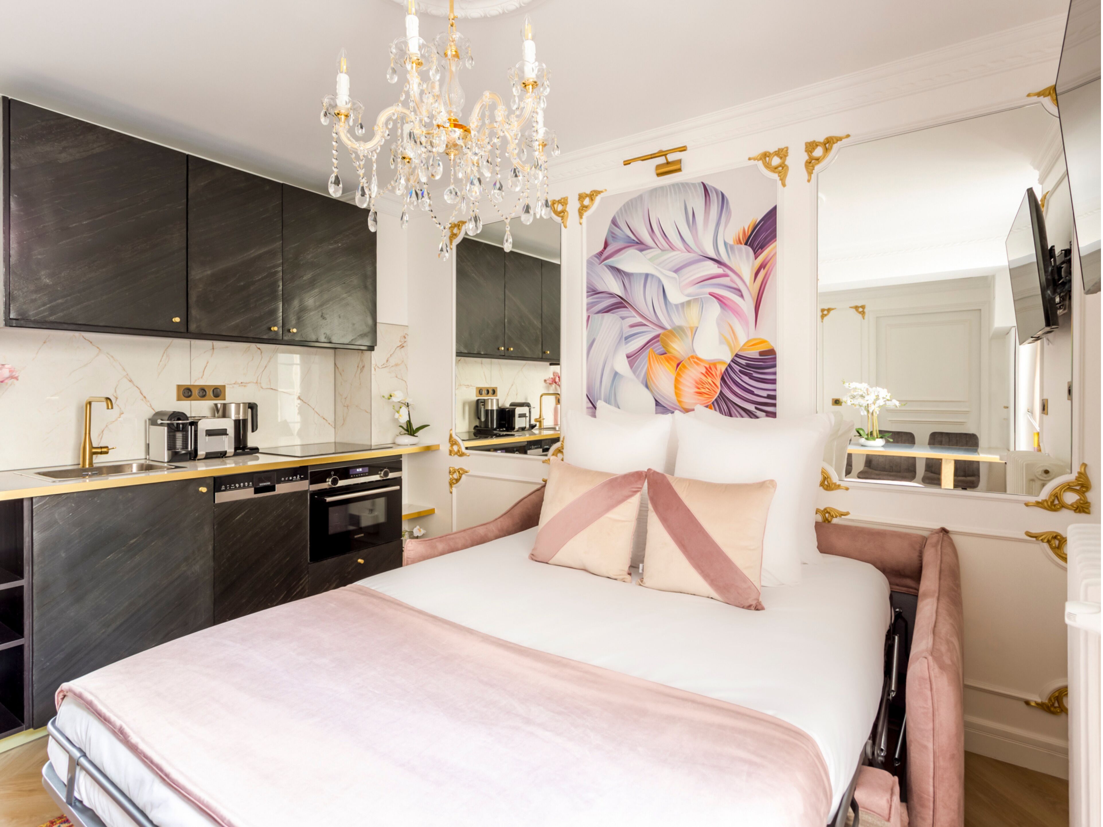 A modern bedroom with a large bed, a chandelier, a painting, and a kitchen area.