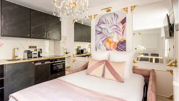A modern bedroom with a large bed, a chandelier, a painting, and a kitchen area.