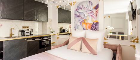 A modern bedroom with a large bed, a chandelier, a painting, and a kitchen area.