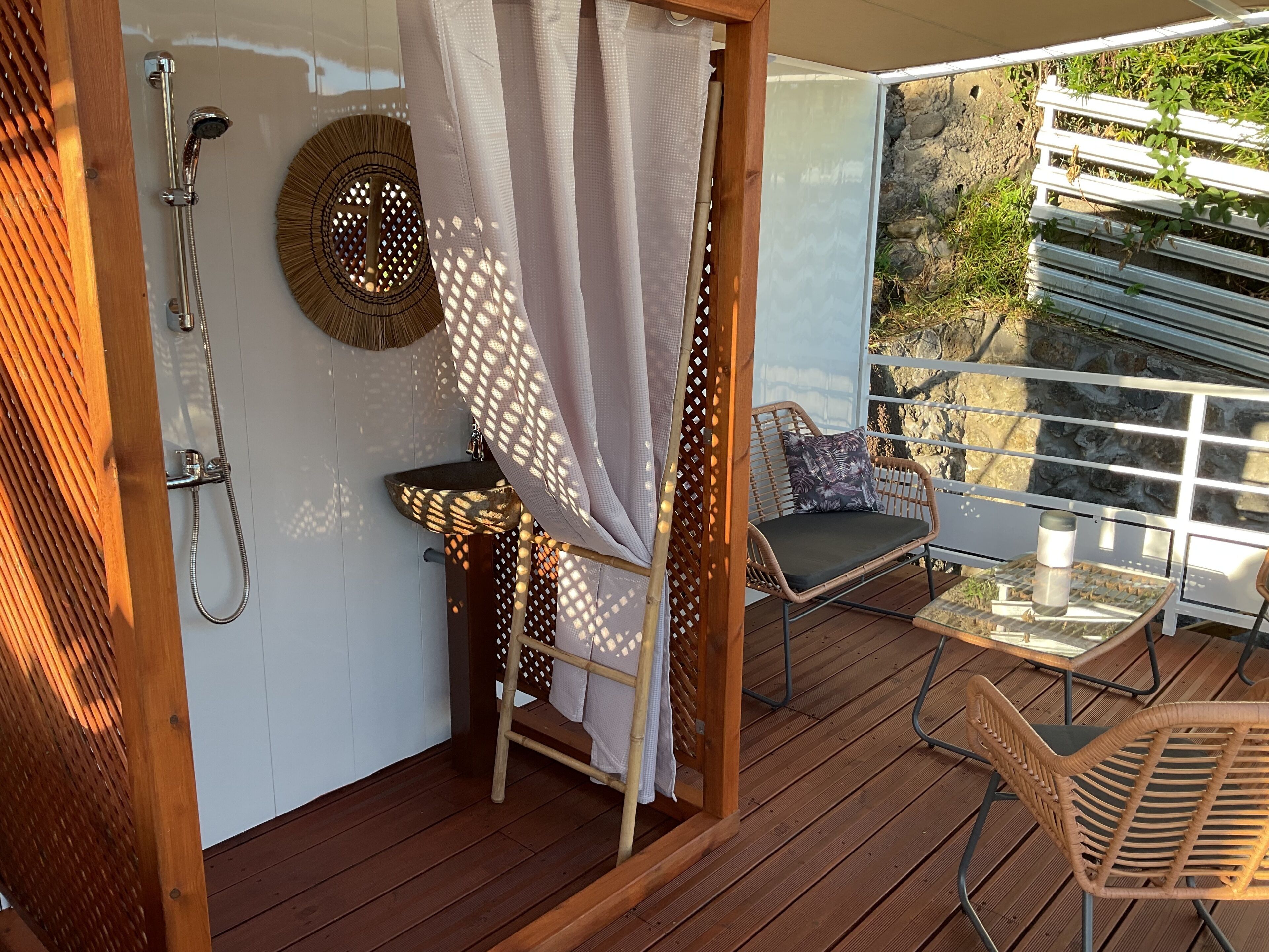 A wooden deck with a shower, a wicker chair, and a table with a metal cup.