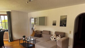 A living room with a sectional sofa, a glass coffee table, and a view through a window with curtains.