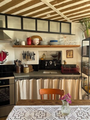 A kitchen with wooden shelves, a sink, and a dining table with a lace tablecloth. - Le Petit Château is a village house in the charming village of Goult. (Goult)