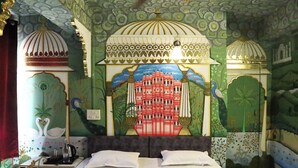 Habitación de hotel con cama, escritorio y un mural que muestra un edificio y pavos reales.