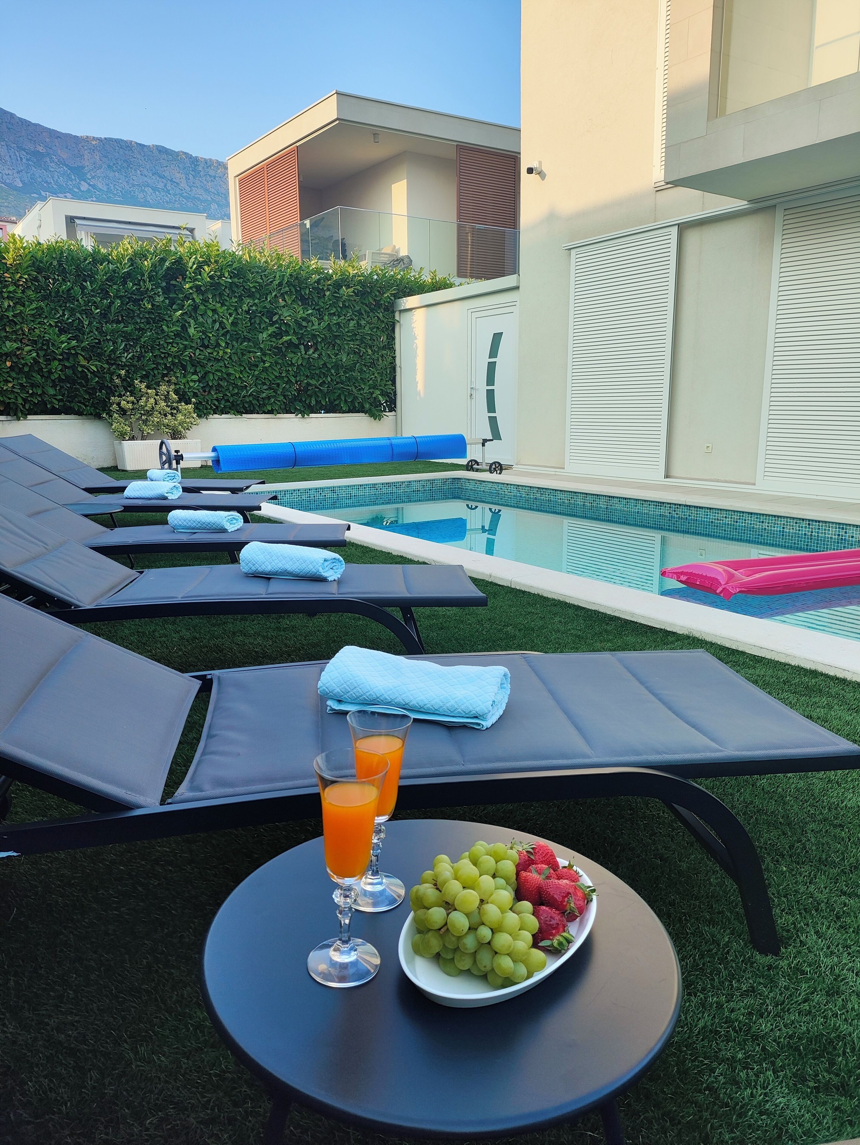 A poolside area with lounge chairs, a table with a fruit bowl and drinks, and a view of a mountain in the background.