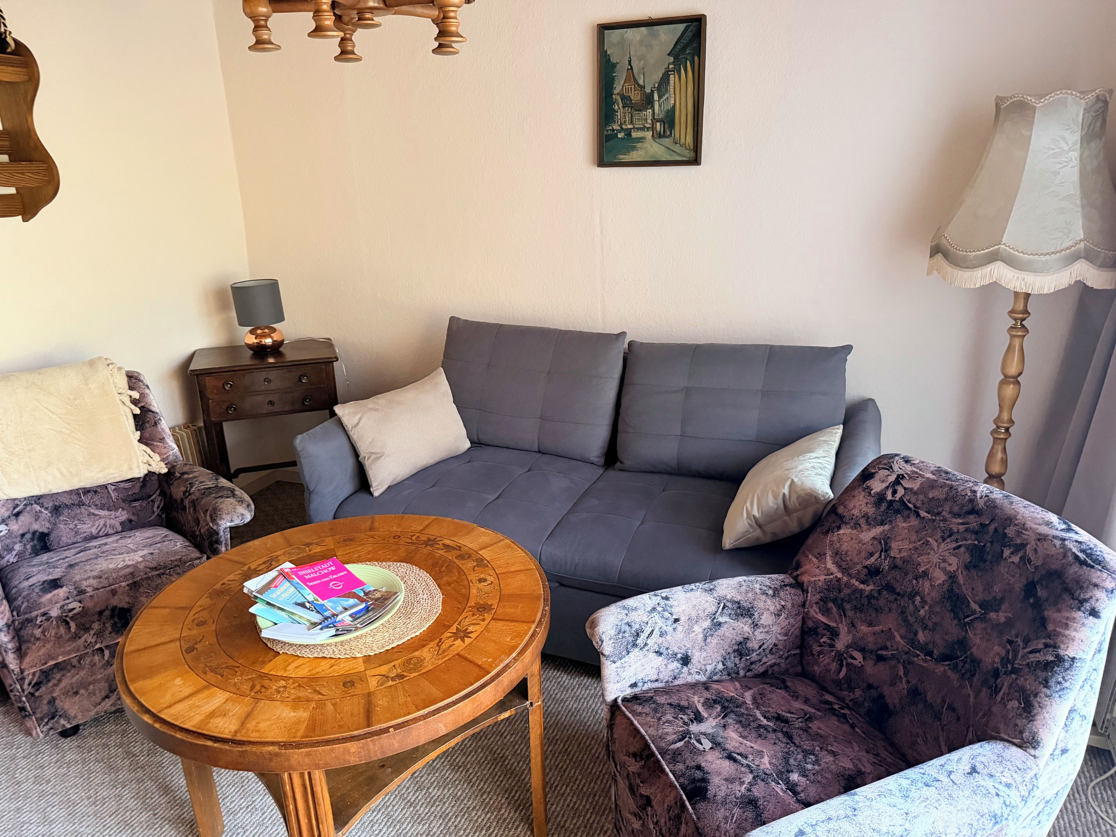 Een woonkamer met een grijze bank, twee fauteuils met een patroon, een houten salontafel met tijdschriften, een lamp en een schilderij aan de muur.
