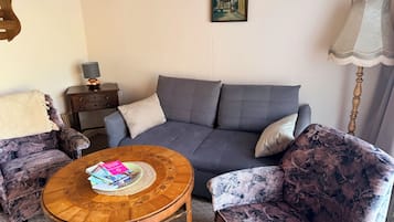 Een woonkamer met een grijze bank, twee fauteuils met een patroon, een houten salontafel met tijdschriften, een lamp en een schilderij aan de muur.