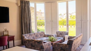 A room with a sofa set, a coffee table, a TV, and a view of a garden through large windows. - Poets 24 Apartments (Ely)