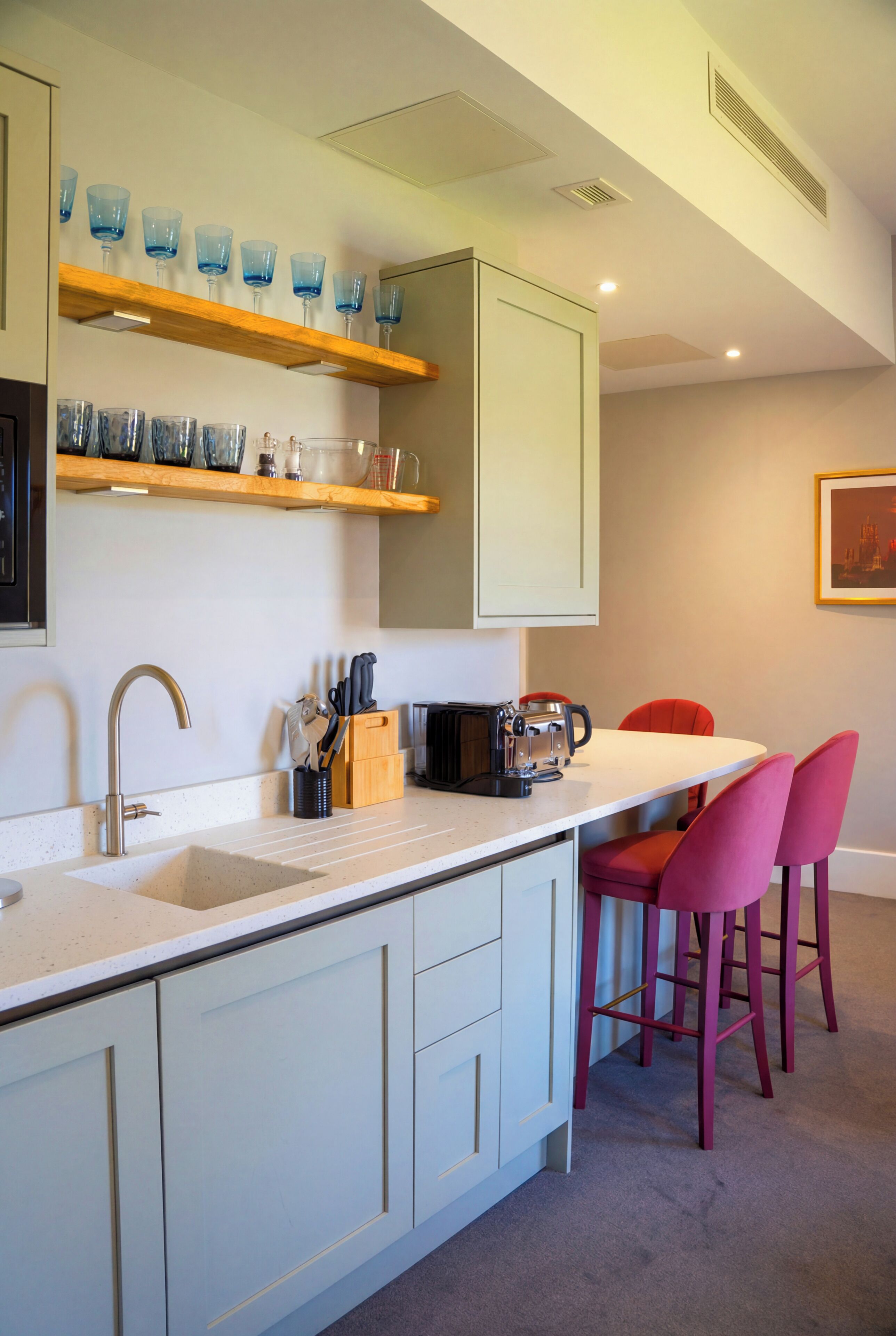 A kitchen with a white countertop, a sink, a coffee maker, and a toaster. There are two red chairs and a wooden shelf with blue glasses.