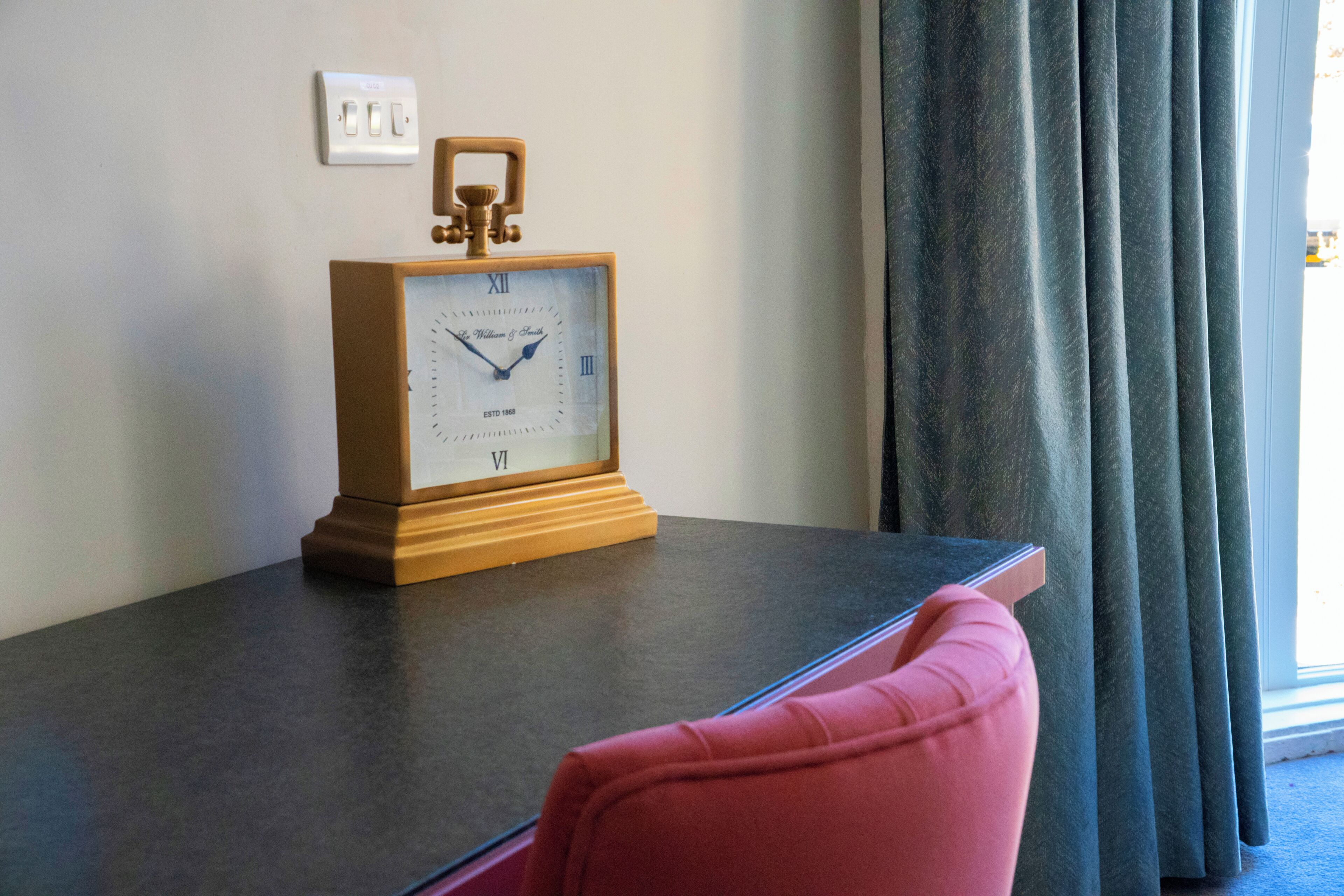 A wooden clock with Roman numerals on a dark surface, a power outlet, and a window with curtains.