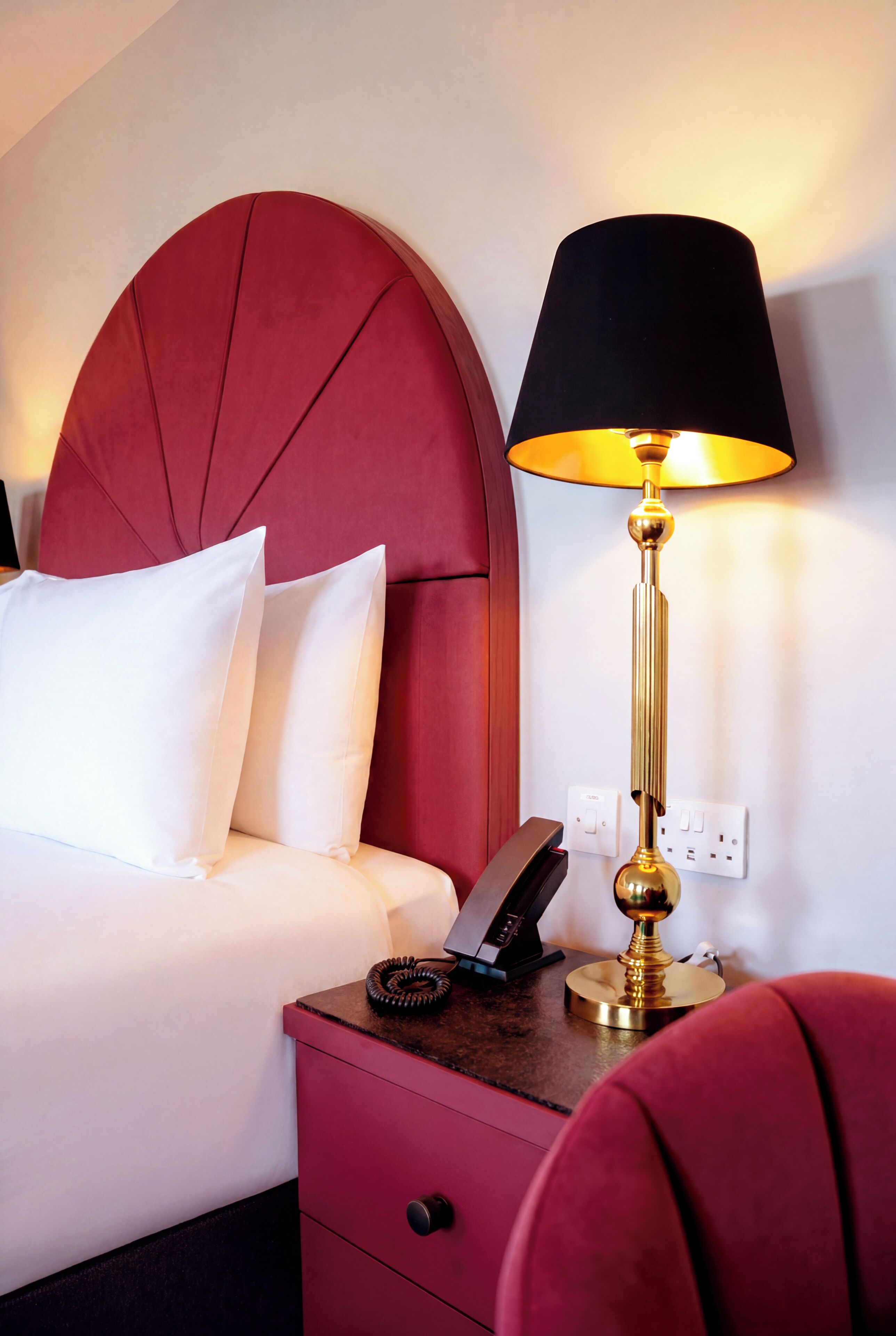 Une chambre d’hôtel avec une tête de lit rembourrée en tissu rouge, une table de chevet avec une lampe et un téléphone.