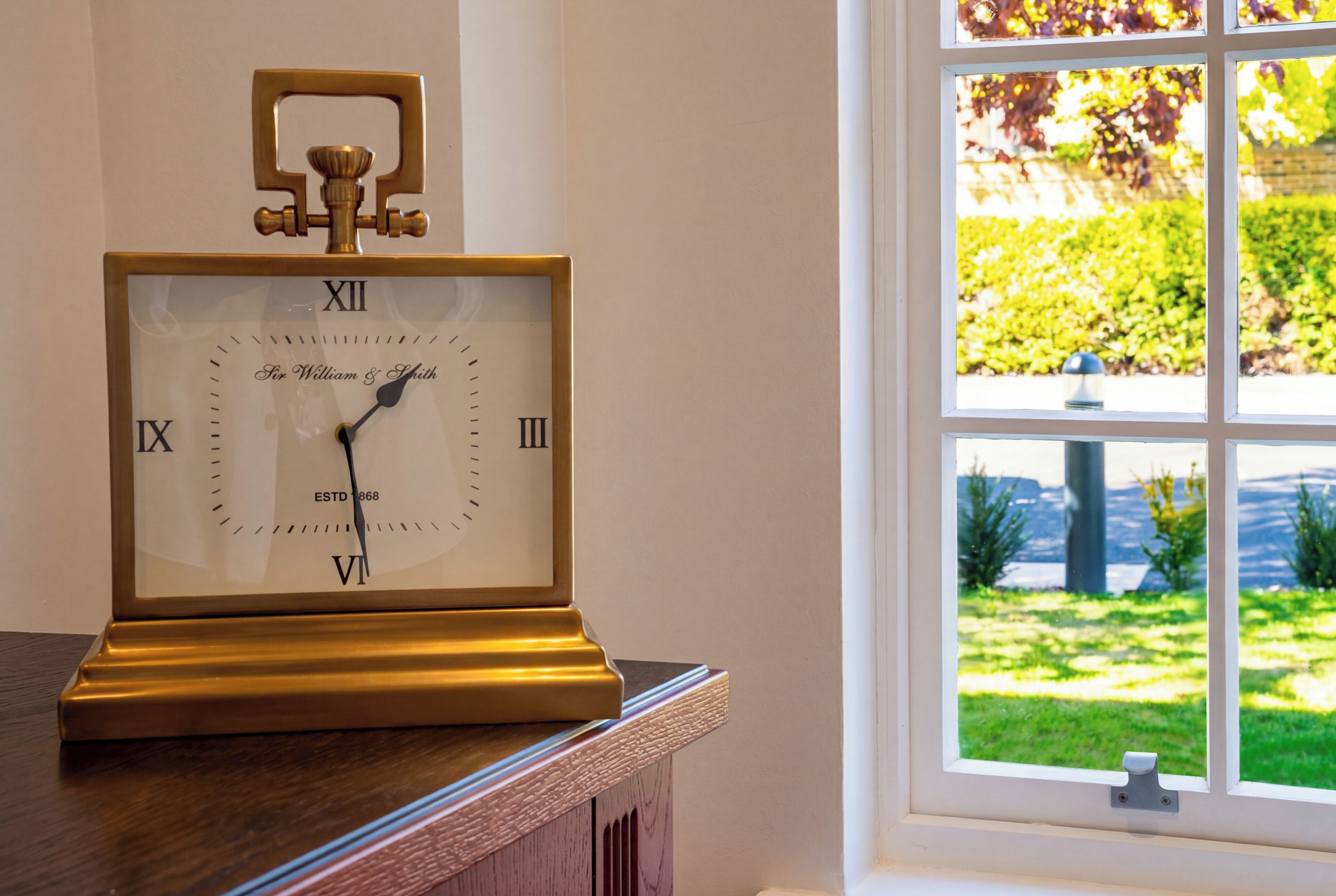 A wall-mounted clock with Roman numerals and a brass pendulum, positioned near a window with a view of greenery.