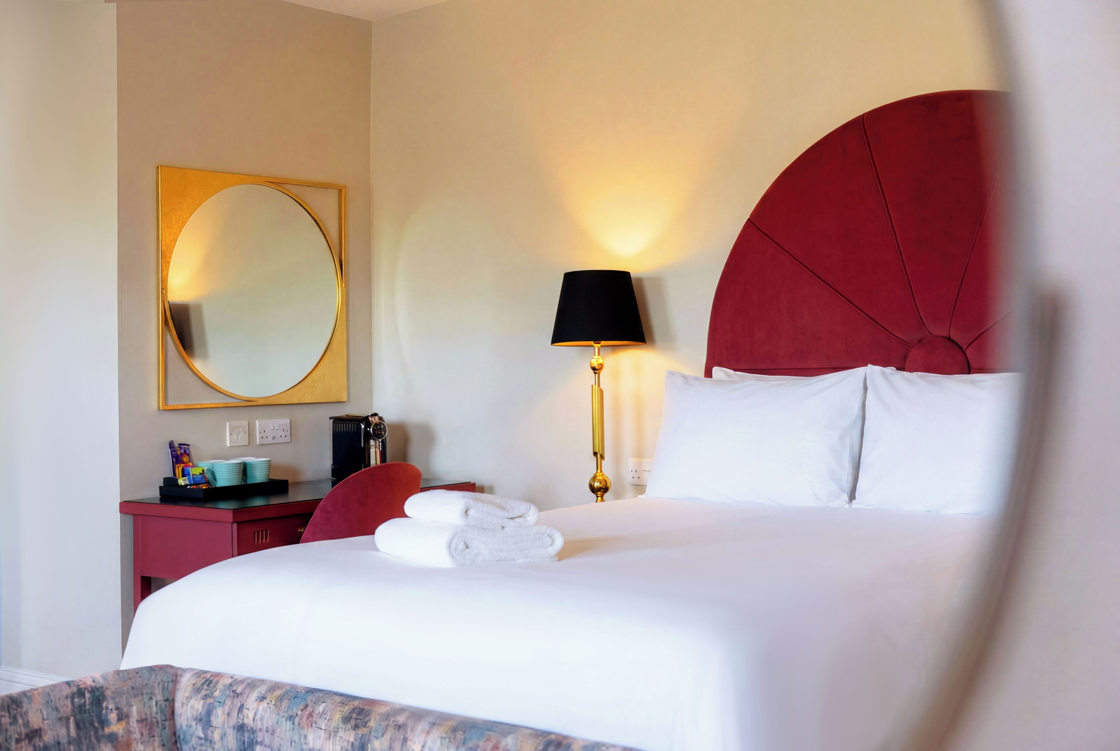 A hotel room with a bed, a desk with a chair, a lamp, a mirror, and a red headboard.
